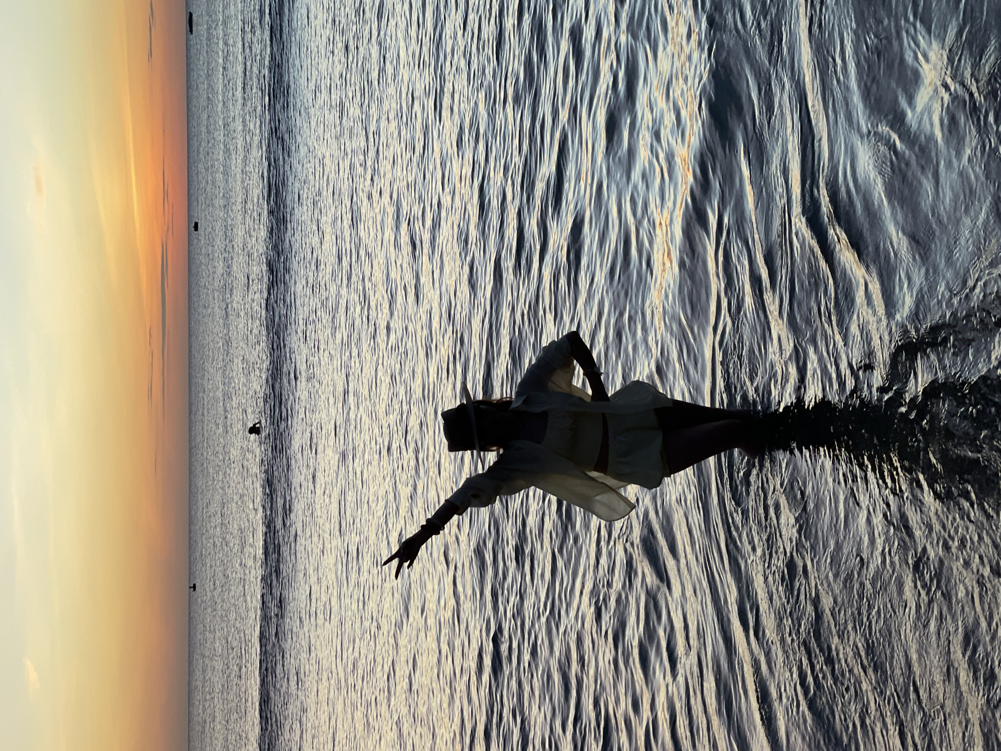 A silhouetted person wearing a hat and a light-colored outfit stands in shallow water at sunset. Their left arm is raised, with the index and middle fingers extended in a peace sign. The person's right hand is on their hip. The water is calm with gentle ripples reflecting the soft colors of the sky. In the background, the ocean stretches to the horizon, where the sky transitions from orange and yellow at the horizon to a lighter blue above. Several small boats and a distant person can be seen on the water. The scene appears to be a beach in Da Nang, Vietnam, likely My Khe Beach, known for its beautiful sunsets.