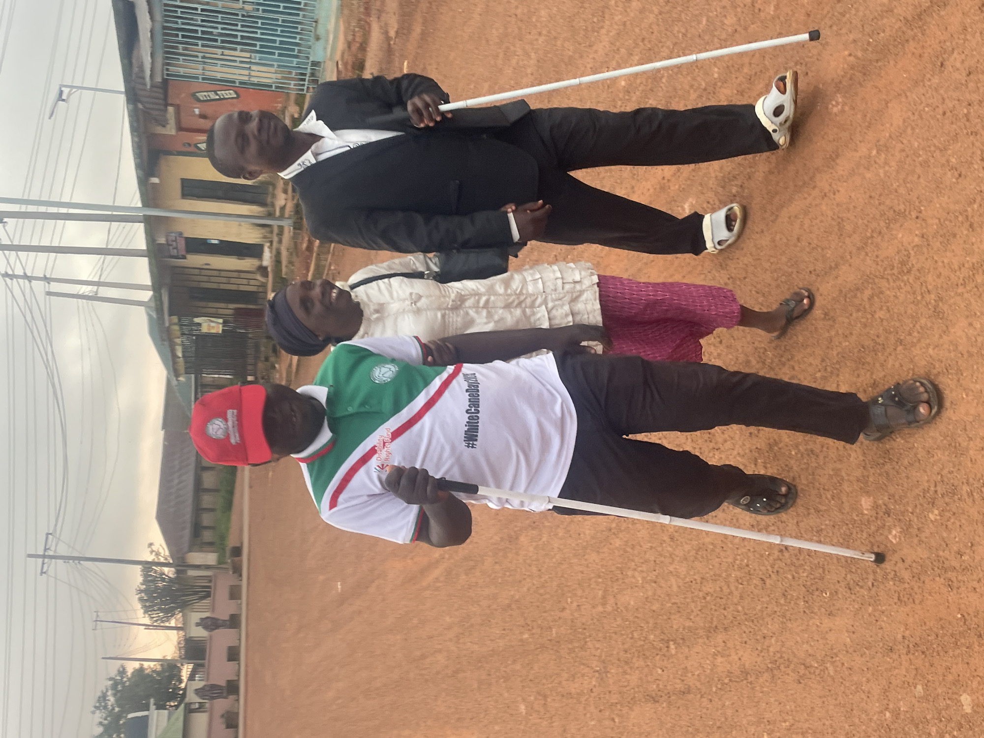 Three people are walking on a dirt road in Gura Top, Nigeria. The person on the left is wearing a red baseball cap, a white polo shirt with green and red trim, and black pants. They are holding a white cane and appear to be visually impaired. The person in the middle is wearing a headscarf, a cream-colored, textured jacket, and pink pants. They are smiling. The person on the right is wearing a black suit jacket, a white shirt, and black pants. They are also holding a white cane and appear to be visually impaired. They are walking in the same direction. The background shows buildings with corrugated metal roofs and utility poles. The sky is overcast.