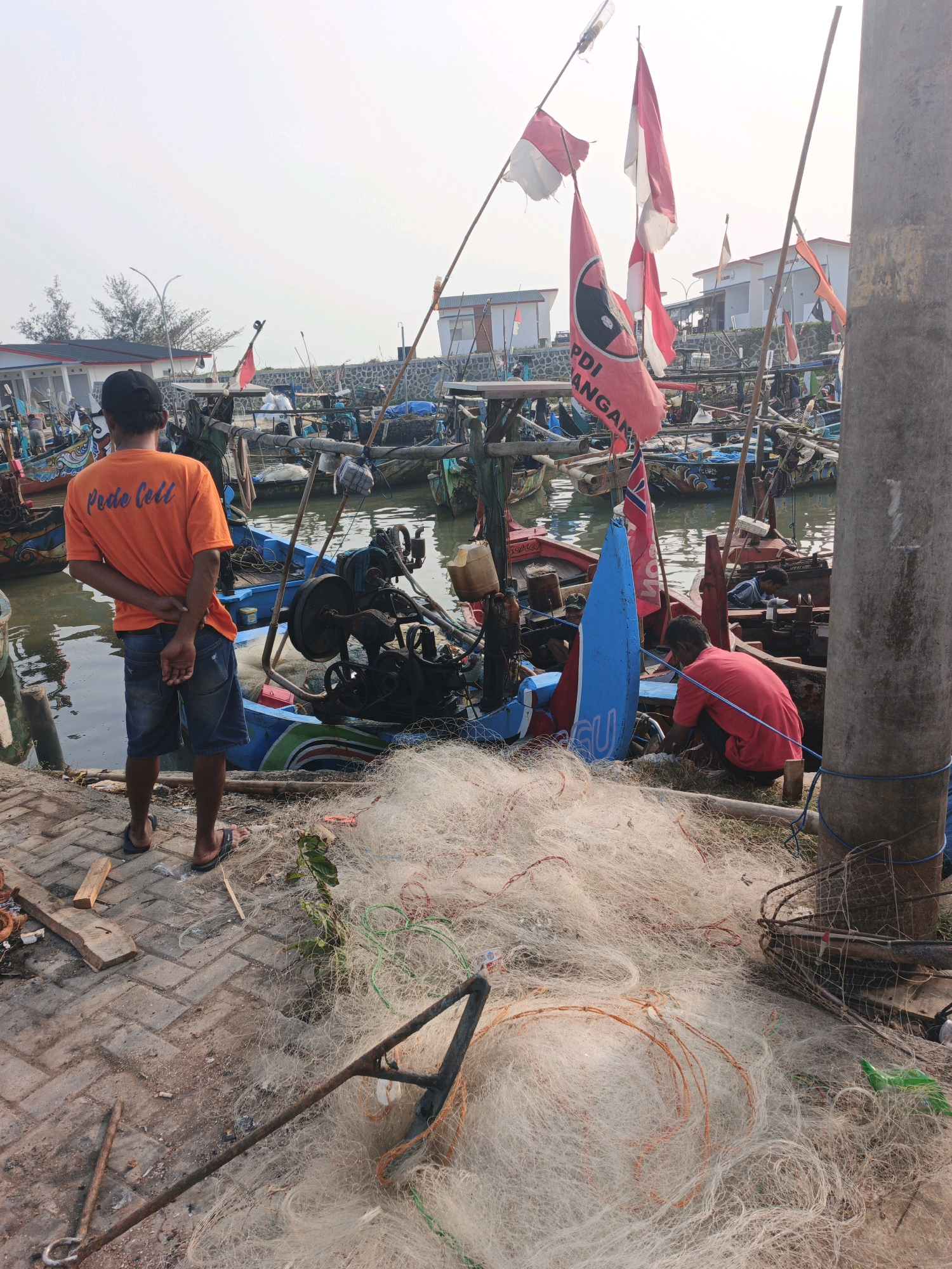 The image captures a harbor scene in Indonesia, possibly a fishing village, with numerous fishing boats docked. In the foreground, a person wearing an orange t-shirt with the text "Pede Cell" and denim shorts stands with their back to the camera. To the right, another person in a red t-shirt is crouching near some fishing nets. A large pile of white fishing nets lies on the ground between them and the boats. Several flags, including Indonesian flags and a red flag with a logo, are visible on poles amidst the boats. The environment appears to be a waterfront with brick paving in the foreground and a gray cylindrical object, likely a pillar, on the right edge of the frame. The sky is bright, suggesting daytime.