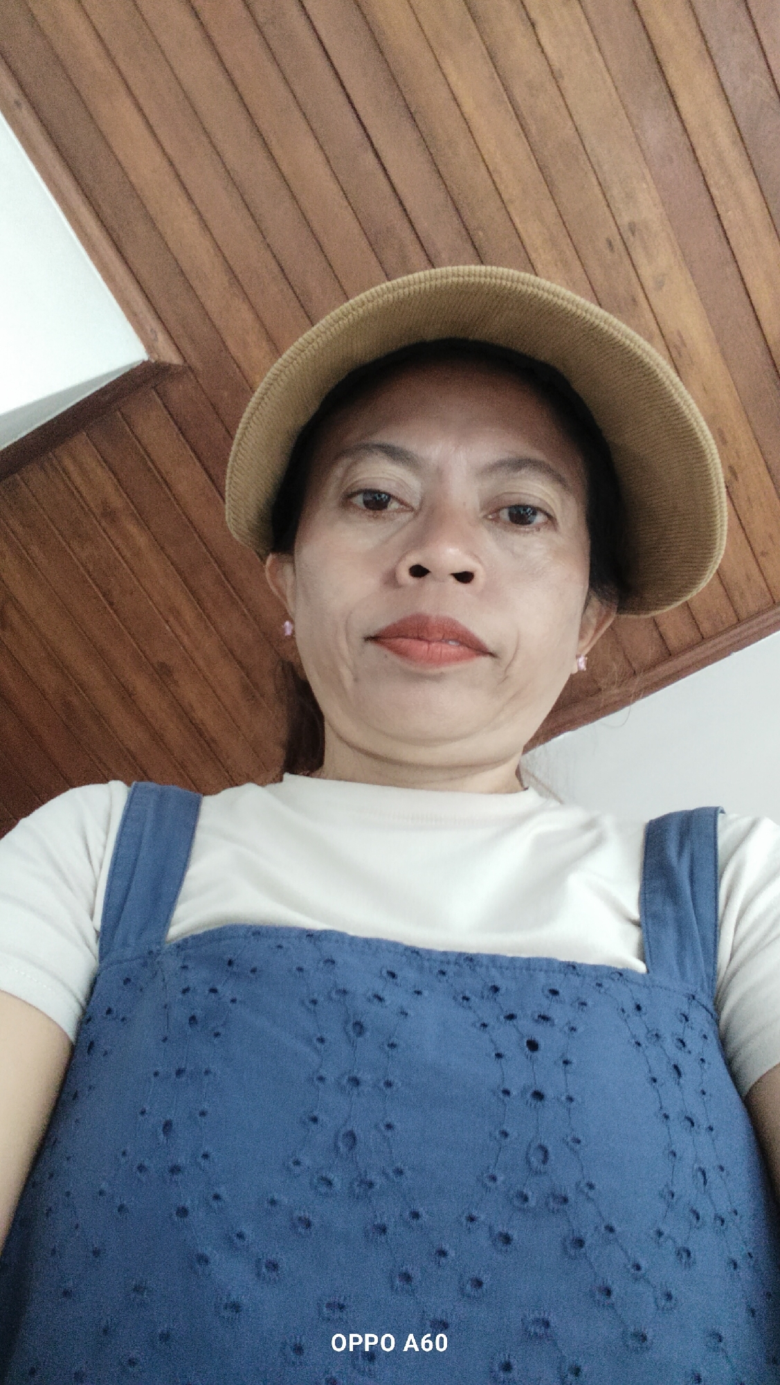 A medium shot, taken from a low angle, shows a woman with dark hair, wearing a tan hat, a white t-shirt, and a blue denim overall dress with eyelet detailing. She has light-colored earrings. Her expression is neutral, and she is looking directly at the camera. The background consists of a wooden ceiling with slatted panels, and a white wall. The image is branded with "OPPO A60" in the bottom right corner, indicating it was taken with an Oppo A60 smartphone. The setting appears to be indoors, likely a home or a casual establishment in North Kuta, Indonesia.