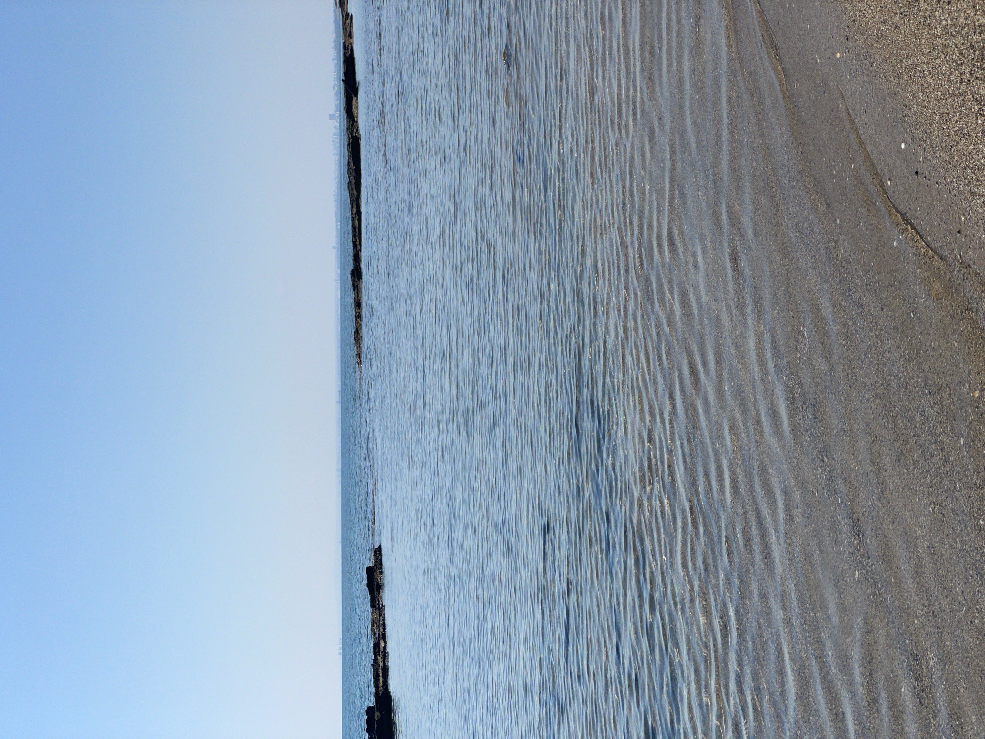 A calm, clear blue sea stretches out under a bright, cloudless sky. In the foreground, a sandy beach slopes gently into the water. The shallow water creates rippled patterns on the sandy seabed, which is visible through the translucent surface. A line of dark rocks, partially submerged, breaks the surface of the water in the middle distance. Beyond the rocks, a faint, hazy line of buildings suggests a distant coastline or city. The scene is serene and empty of people, evoking a sense of quiet solitude.