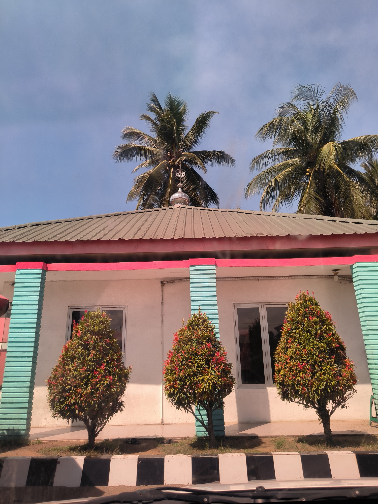 The image displays the exterior of a small building, likely a mosque or prayer house, beneath a bright blue sky. The building features white walls with distinct turquoise and pink trim, including prominent, fluted turquoise columns. It is topped with a green corrugated metal roof, from which a small, silver-colored dome ascends. A finial, bearing the word "Allah" in stylized Arabic script, crowns the dome.
Behind the building, several tall palm trees stand, their fronds fanning out against the clear sky, indicating a tropical environment. In front of the building's two visible windows, three conical bushes with vibrant green and reddish-orange leaves are neatly arranged.
The photograph was taken during bright daylight, under sunny conditions with minimal cloud cover. The foreground reveals a black and white checkered curb or barrier, with the edge of a vehicle's dashboard visible, suggesting the photo was captured from inside a car. The scene conveys a sense of calm in Cotpuuk, Indonesia.