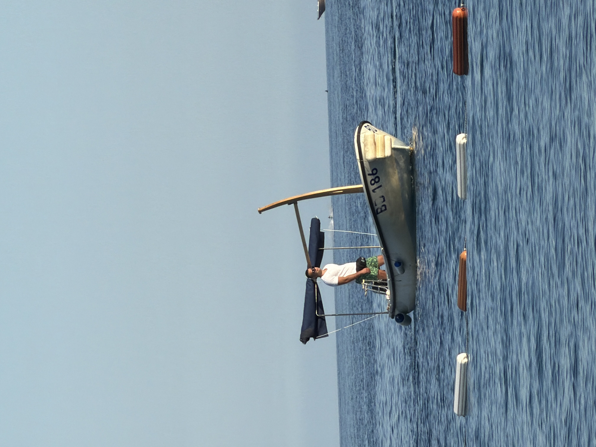 The image shows a man on a sailboat in the water, possibly off the coast of Budva, Montenegro. The boat has the identification number "BD 186". The man is wearing a white shirt and patterned shorts. There are buoys in the water near the boat. The sky is light blue.