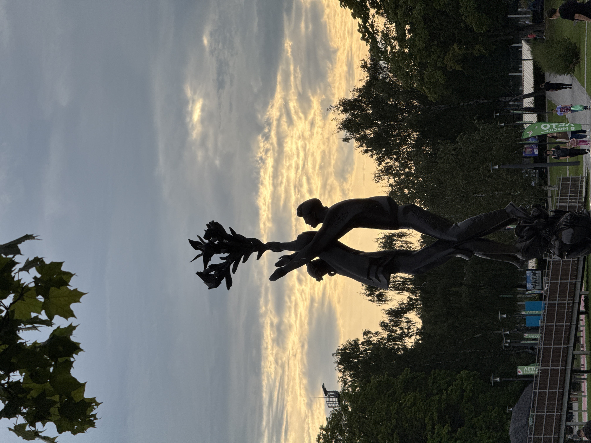 The image depicts a bronze sculpture of three figures reaching upwards, holding a branch with leaves and birds. The sculpture is silhouetted against a cloudy sky with streaks of yellow and orange, suggesting either sunrise or sunset. In the background, there are trees, lampposts, a pedestrian bridge, and a few indistinct figures walking. A tall structure, possibly a tower, is visible in the distance on the left. The overall scene appears to be an outdoor public space in Khimki, Russia, likely a park or plaza, given the presence of the sculpture, greenery, and pedestrian activity. The lighting conditions indicate it is either early morning or late evening.