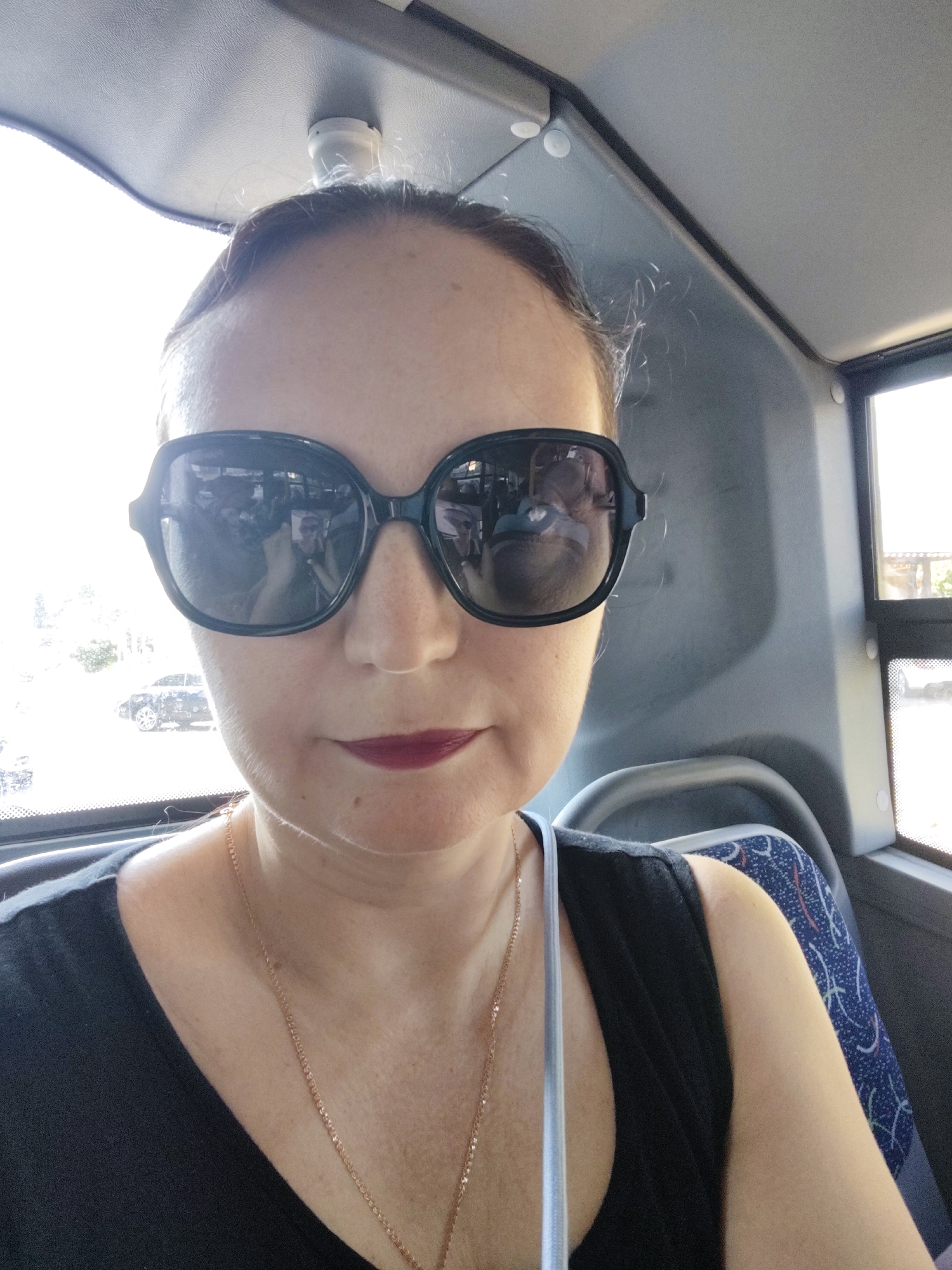 In a close-up selfie taken from inside a vehicle, a woman with fair skin and dark hair pulled back wears oversized black sunglasses and dark lipstick. Her gaze is directed towards the camera, and a portion of her shoulders and upper chest is visible. She is dressed in a black top and a thin gold necklace. Reflections in her sunglasses show people inside the vehicle and a blurred exterior view of a dark car. The background suggests the interior of a bus or similar public transport, with a grey headliner and seats visible. The image appears to have been taken during the daytime, with bright light entering from the side windows. The location is identified as Sochi, Russia.