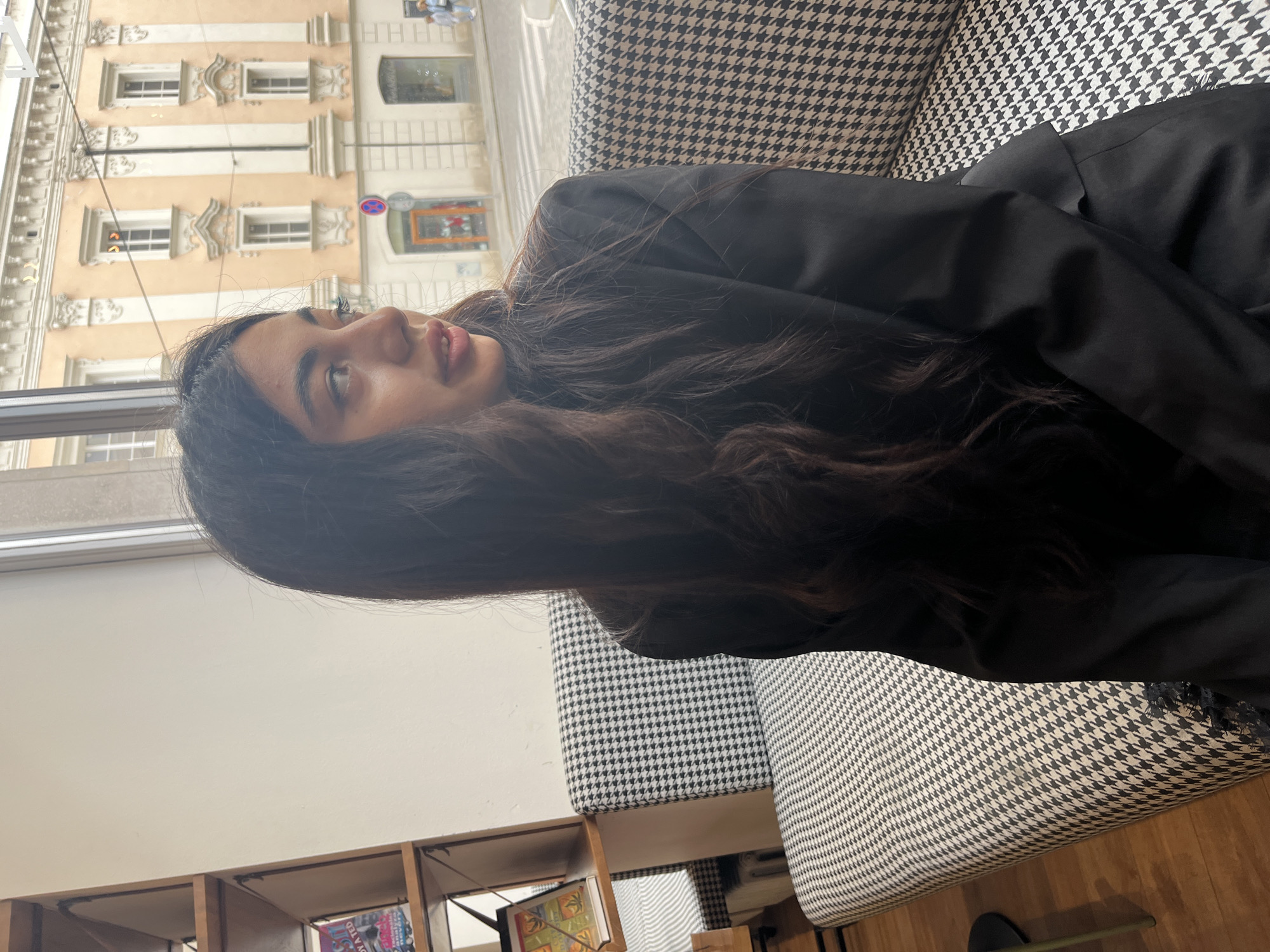 A young woman with long, dark, wavy hair sits on a bench with a black and white houndstooth pattern in Pilsen, Czechia. She is wearing a black blazer and is looking to the right. Behind her, through a window, is a street scene with an ornate building and a crosswalk. To the left of the woman is a bookshelf with some books. The overall setting appears to be a cafe or a library.
