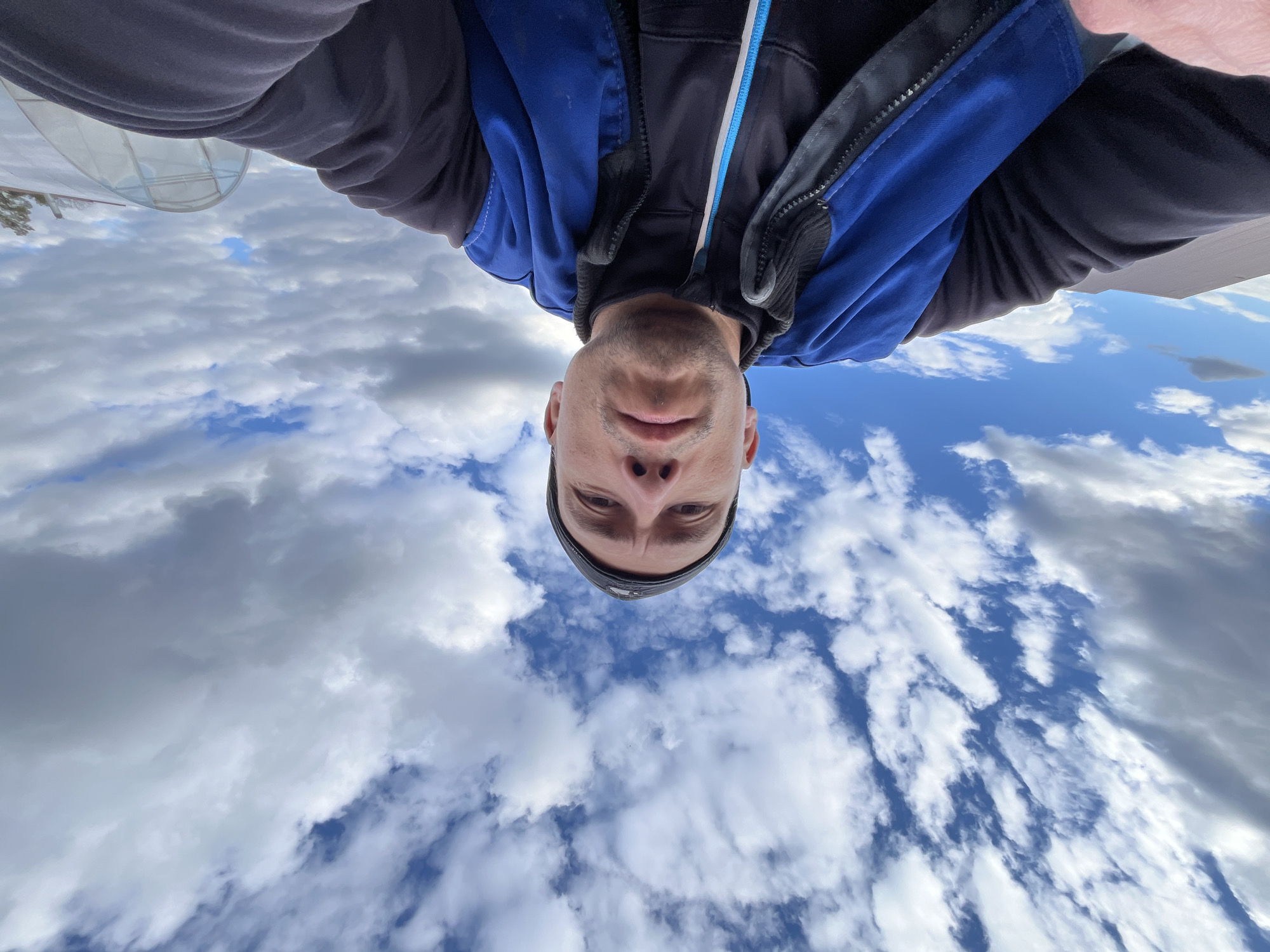 A low-angle selfie photograph depicts a man with a fair complexion, a close-cropped haircut, and a dark beanie, positioned against a backdrop of a partly cloudy sky with some areas of clear blue. He is wearing a dark hooded sweatshirt underneath a blue and black jacket. Some type of transparent greenhouse-type structure is also visible near the top left of the photograph. There is no evidence to confirm the image was taken in Chaikovsky, Russia.