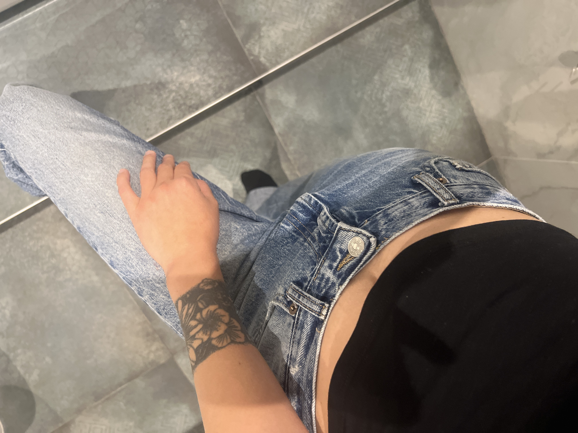 A high-angle shot depicts a person wearing light blue jeans and a black top, standing on a gray tile floor. The person's left arm, featuring a floral tattoo on the wrist, rests on their left leg. A black sock is visible on the right foot. The environment suggests an indoor setting, possibly a bathroom or similar tiled area.