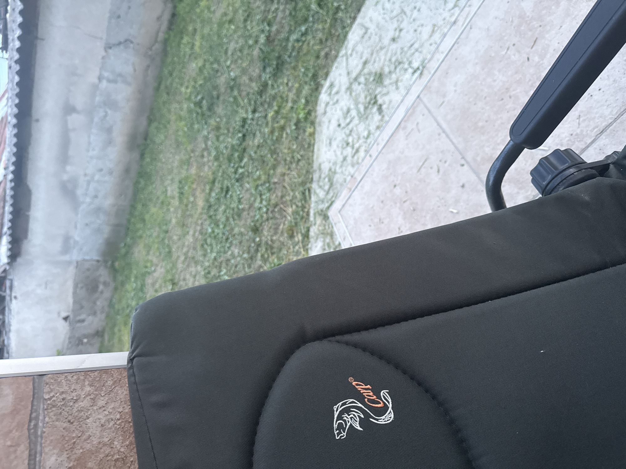 This image, taken in Rogosh, Bulgaria, depicts an outdoor scene focusing on a dark grey or black upholstered chair. The chair features an embroidered white outline of a jumping fish (carp) with the word "Carp" in orange script and a registered trademark symbol (®) positioned below it on the left side of the backrest. A black armrest or support frame with a circular adjustment knob is visible in the bottom right corner of the chair. The chair rests on light-colored tiled flooring, which is scattered with numerous small, dry clippings, likely grass. Beyond the tiled area, a patch of green grass, also interspersed with clippings, occupies the midground. The background is dominated by a light grey concrete wall exhibiting textural variations and weathering. Above the wall, a light-colored corrugated roof with a scalloped edge is visible. A section of reddish-brown brickwork is present in the far upper-left background. No individuals or active subjects are present in the scene, which is illuminated by natural daylight.