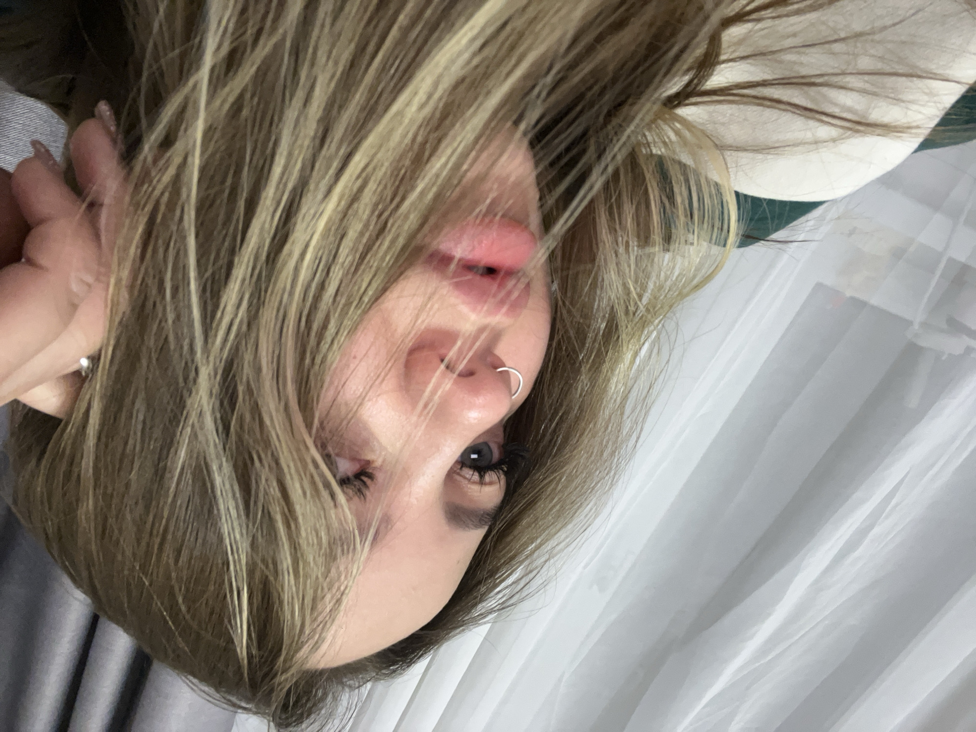 A selfie of a woman indoors. She has long, blonde hair and fair skin. Her left eye is visible, with dark eyelashes. She has a silver nose ring. Her lips are pink. Her left hand is raised near her face, with a silver ring visible on one finger. The background includes a white sheer curtain and a glimpse of a white and green garment. The location is indeterminate; there is no evidence in the image to place the location in Krasnodar, Russia.