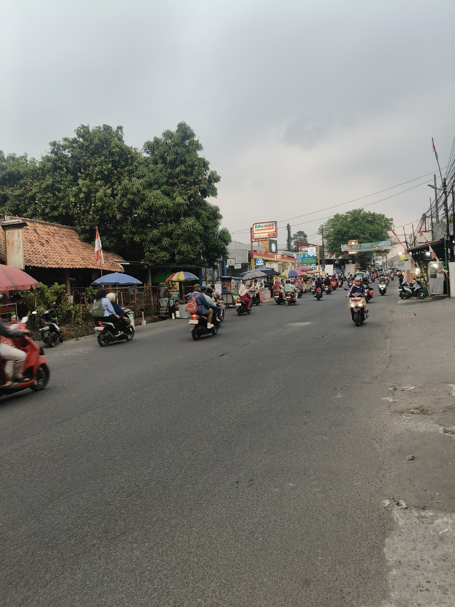 This image captures a busy street scene in South Tangerang, Indonesia. A multitude of motorcycles fills the asphalt road, creating a sense of motion blur. Several riders are visible, some with passengers, navigating the traffic. On the left side of the road, there's a building with a traditional tiled roof and an Indonesian flag. Adjacent to it are several market stalls with colorful umbrellas, selling various items. Further down the street, a prominent "Alfamart" convenience store sign is visible, along with other shops and businesses. Power lines crisscross overhead against a cloudy sky. The overall environment appears to be a commercial and residential area with a moderate level of urban activity.