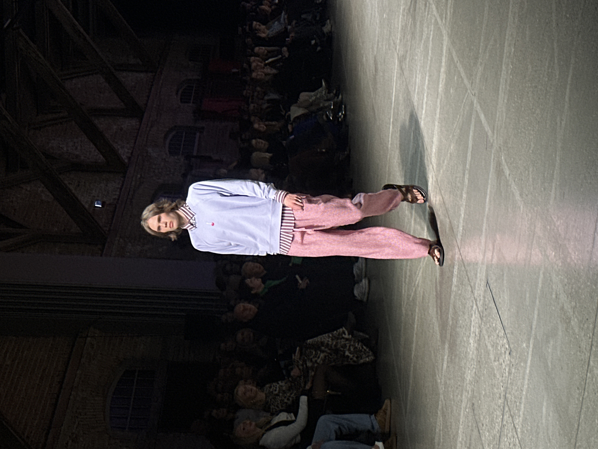 A man walks the runway in a fashion show. He wears a long-sleeved light blue shirt, pink trousers with stripes at the cuffs, and sandals. The audience watches from the sides of the runway. The location appears to be an older building, possibly in Riga, Latvia, based on the style of architecture visible in the background. The runway surface is a light grey material.