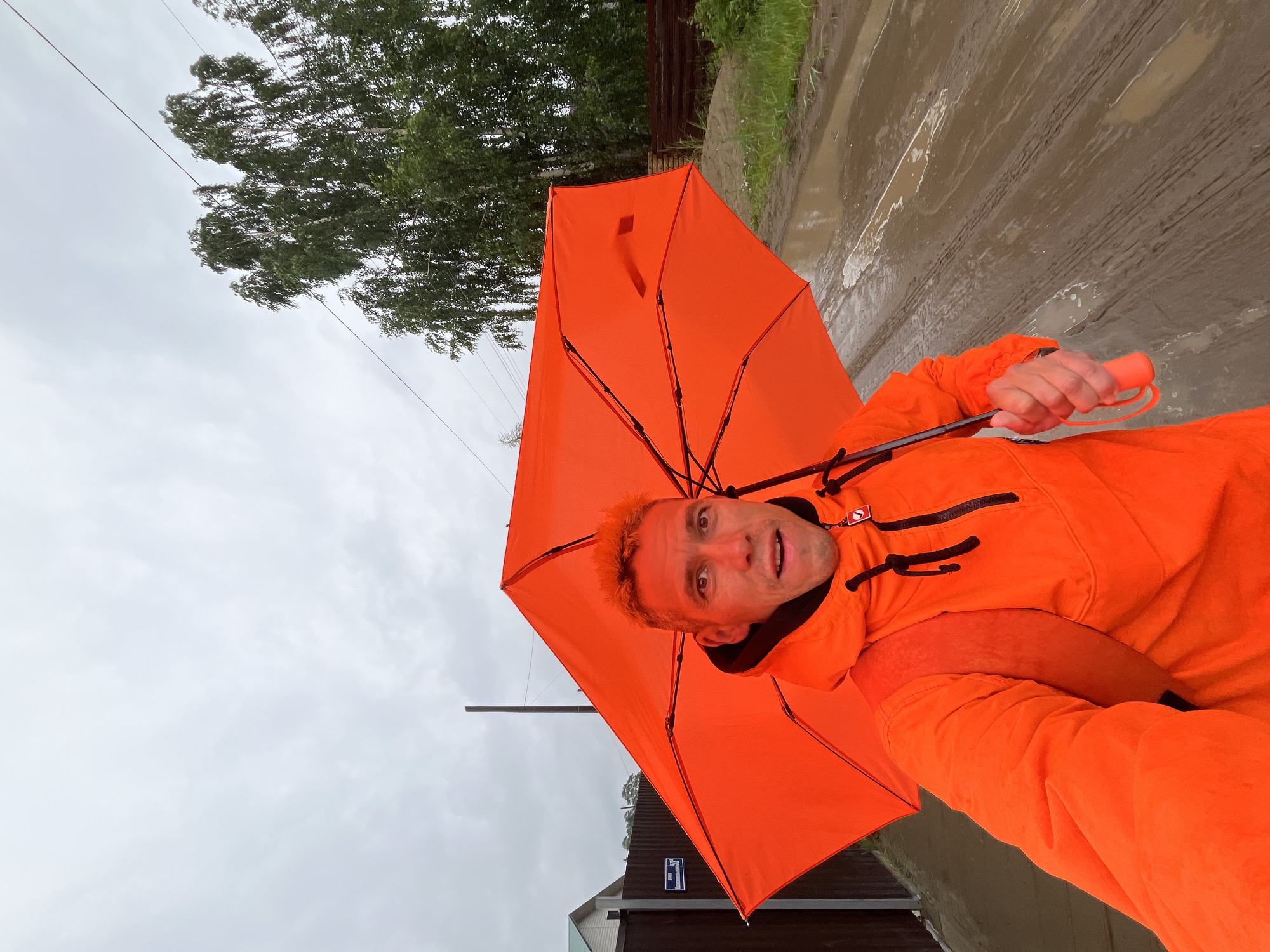 A selfie of a man holding an open orange umbrella under a cloudy sky. He is wearing an orange hooded jacket and a backpack. The man is standing on a muddy road with puddles and tire tracks. In the background, there is a wooden fence, a house, and trees. There are power lines visible in the sky. The location is the city of Chaikovsky, Russia.