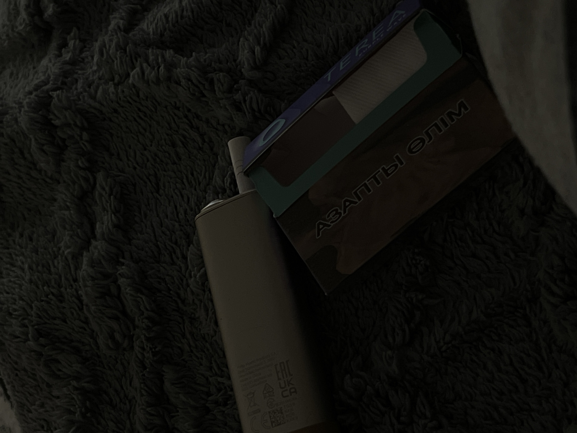 This media file depicts a close-up, dimly lit scene featuring a small, cylindrical electronic device and a box with text. The objects are resting on a dark, textured surface that appears to be a soft blanket or fabric.
The electronic device is light-colored and has some visible text and symbols towards its base, including an "EAC" logo, "UK CA" logos, and what seems to be a model number or serial code. The box is darker and angled diagonally, with text visible on its surface. Part of the text on the box is in a Latin-based alphabet, spelling out "TEREA PURPLE WAVE," and another section appears to be in Cyrillic, reading "АЗАПТЫ ƏЛІМ".
The lighting is low, suggesting it might be nighttime or an indoor setting with subdued illumination. The overall mood is dark and somewhat mysterious due to the poor lighting conditions. There are no people visible in the frame. The location context provided is "Байкент, Kazakhstan," which adds a geographical detail to the scene. The activity taking place is not discernible, as the objects are static. Notable details include the textured background, the specific branding and text on the box and device, and the low-light environment.