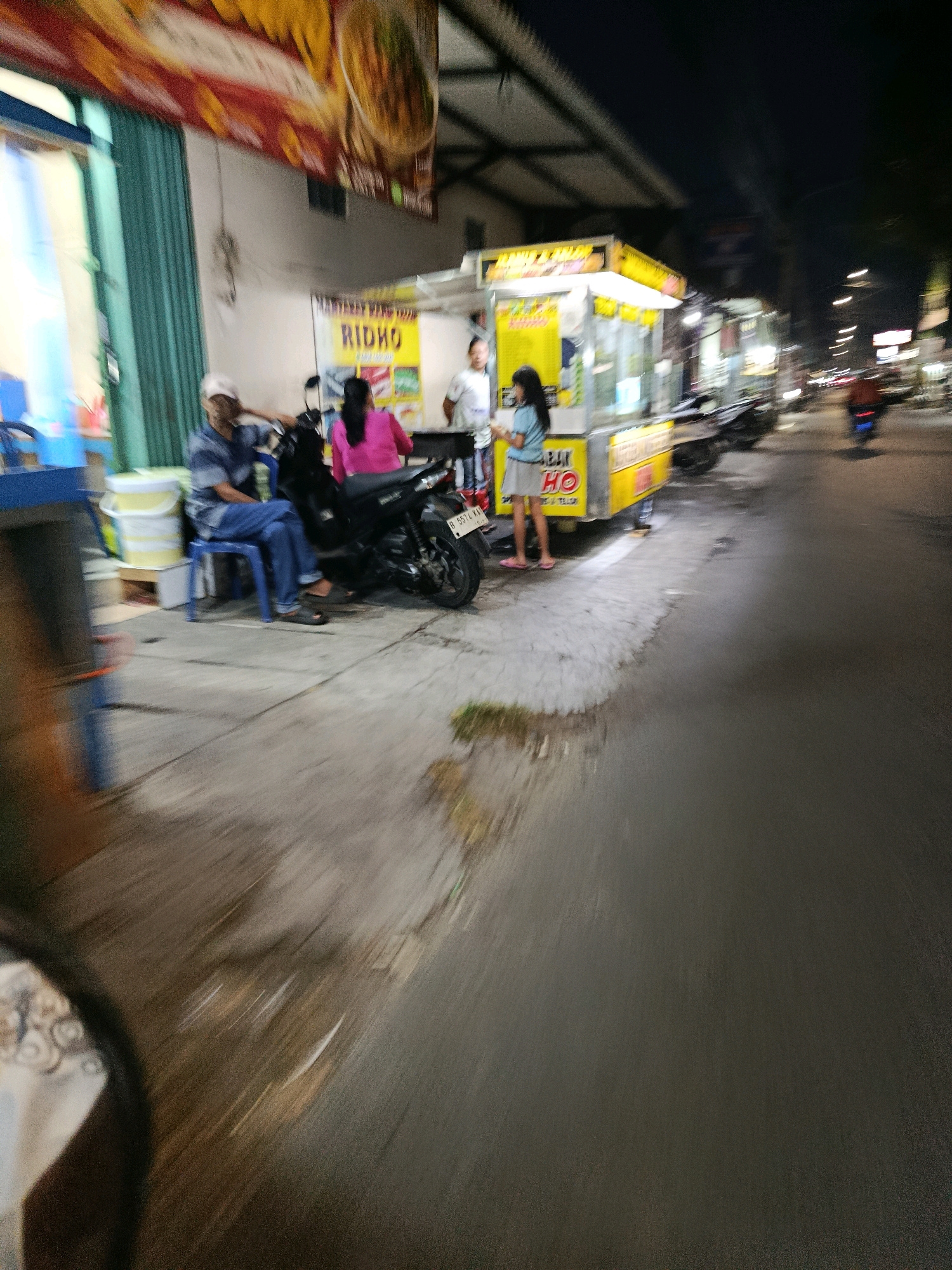 This is a nighttime photograph taken on a street in Bekasi, Indonesia, depicting a food stall scene. A man wearing a blue and white striped shirt and blue jeans sits on a blue plastic chair next to a black motorcycle. A woman in a pink shirt stands behind the motorcycle, facing the food stall. Another woman, appearing to be a customer, stands at the counter of the yellow food stall, which displays the name "RIDHO" in large yellow letters. The stall has a bright yellow awning and appears to be selling food. The street is paved, and a portion of it is covered by the food stall and its immediate surroundings. In the background, other stalls and buildings are visible, along with streetlights and the blurred motion of a motorcycle rider on the road, suggesting a busy urban environment. The overall impression is of a lively street food area at night.