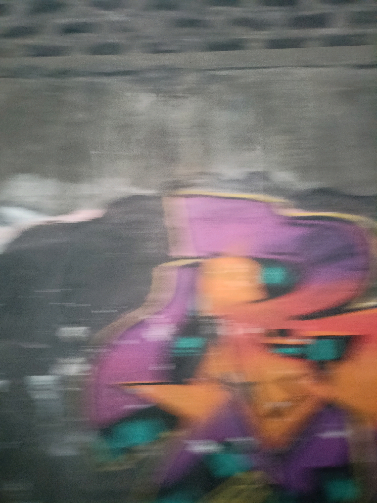 The image depicts a blurry, abstract mural or graffiti on a concrete wall. The mural features large, bold shapes in vibrant colors, primarily purple, orange, and teal. The effect of motion blur suggests the photograph was taken from a moving vehicle. The background appears to be a grey, textured concrete surface, possibly with visible brickwork at the top, indicating an urban environment. The scene is devoid of any people or specific landmarks that would definitively place it in Magelang, Indonesia, although it is characteristic of public art found in urban settings worldwide.