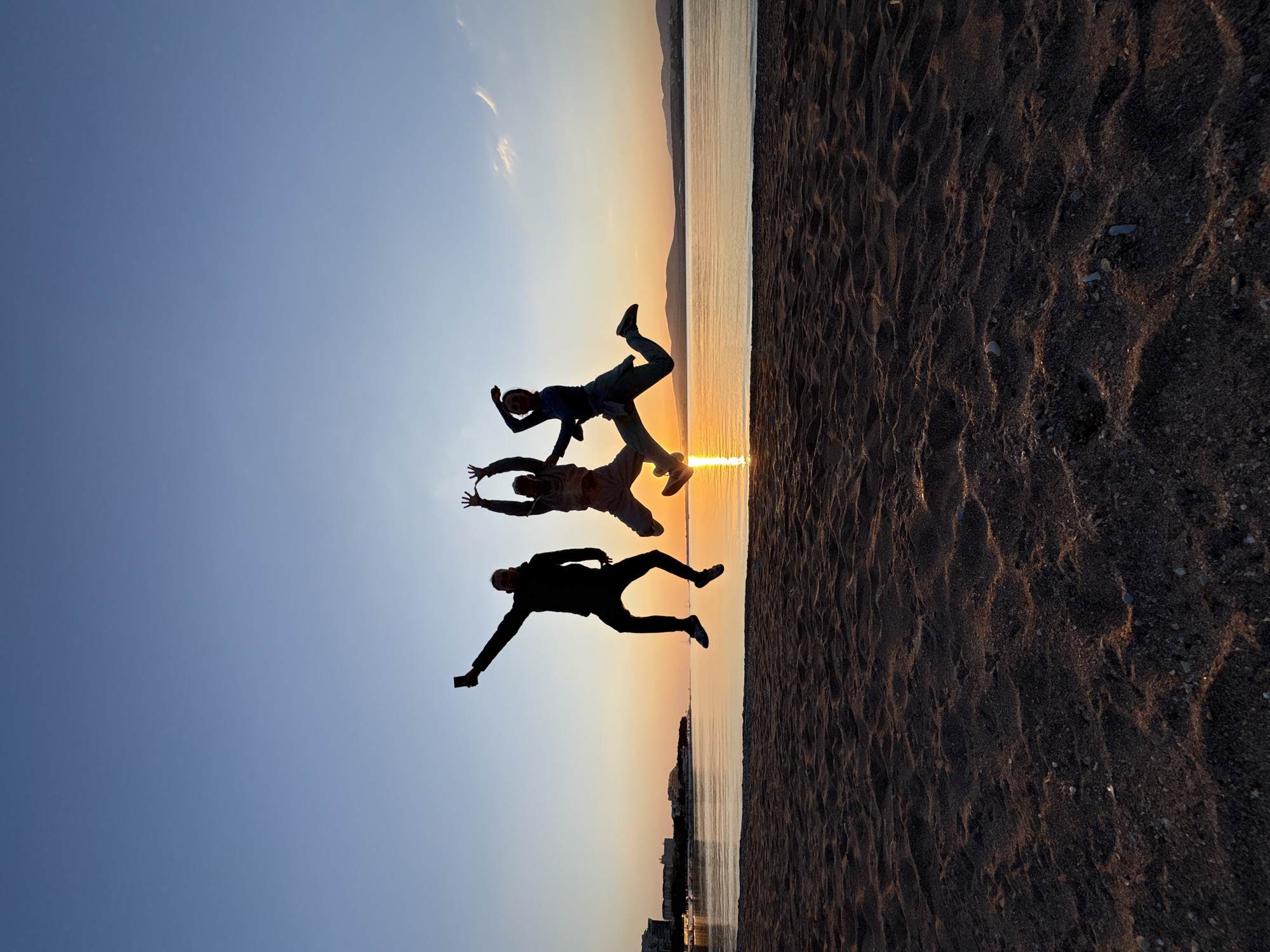 The image depicts a vibrant sunset scene over a body of water in Gelendzhik, Russia, featuring three silhouetted individuals on a beach. The photo is taken at the golden hour, with the sun setting directly over the water, casting a brilliant orange and yellow glow across the horizon and reflecting intensely on the calm surface.
In the foreground, a sandy beach, textured with subtle ripples and small pebbles, occupies the right side of the frame, appearing mostly in shadow. Against the radiant sunset, three people are captured in mid-air, striking dynamic, joyful poses. Their figures are entirely silhouetted, making their actions appear as if they are jumping or floating above the sand. The person on the far left appears to be holding a smartphone in their outstretched hand.
The sky transitions from a deep, clear blue at the top to lighter blues and then to the warm, fiery hues of the sunset at the horizon. A few faint, wispy clouds are visible in the upper left portion of the sky. Across the water, a distant coastline or cityscape is faintly visible against the fading light. The overall mood is one of exuberance, freedom, and warmth, characteristic of an evening on a beach during sunset. There is no visible text in the image.