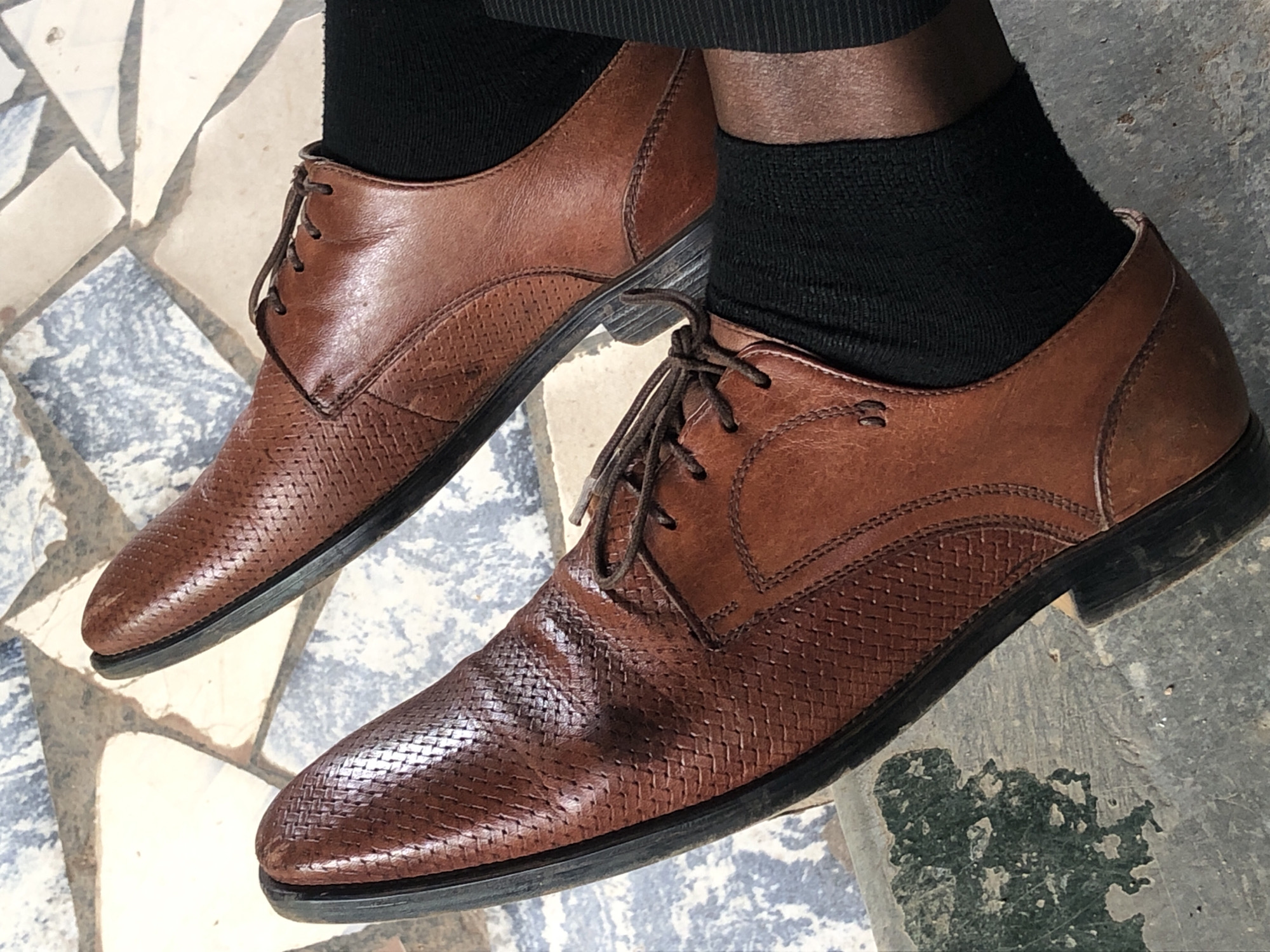 A pair of brown leather oxford shoes with a woven texture and dark brown laces are worn by a person on a mosaic tiled surface. The shoes are made of leather with a glossy finish and are laced up. The person is wearing black socks. The background is a mosaic of geometric tiles in shades of blue, white, and beige. The lighting suggests it is daytime and outdoors.