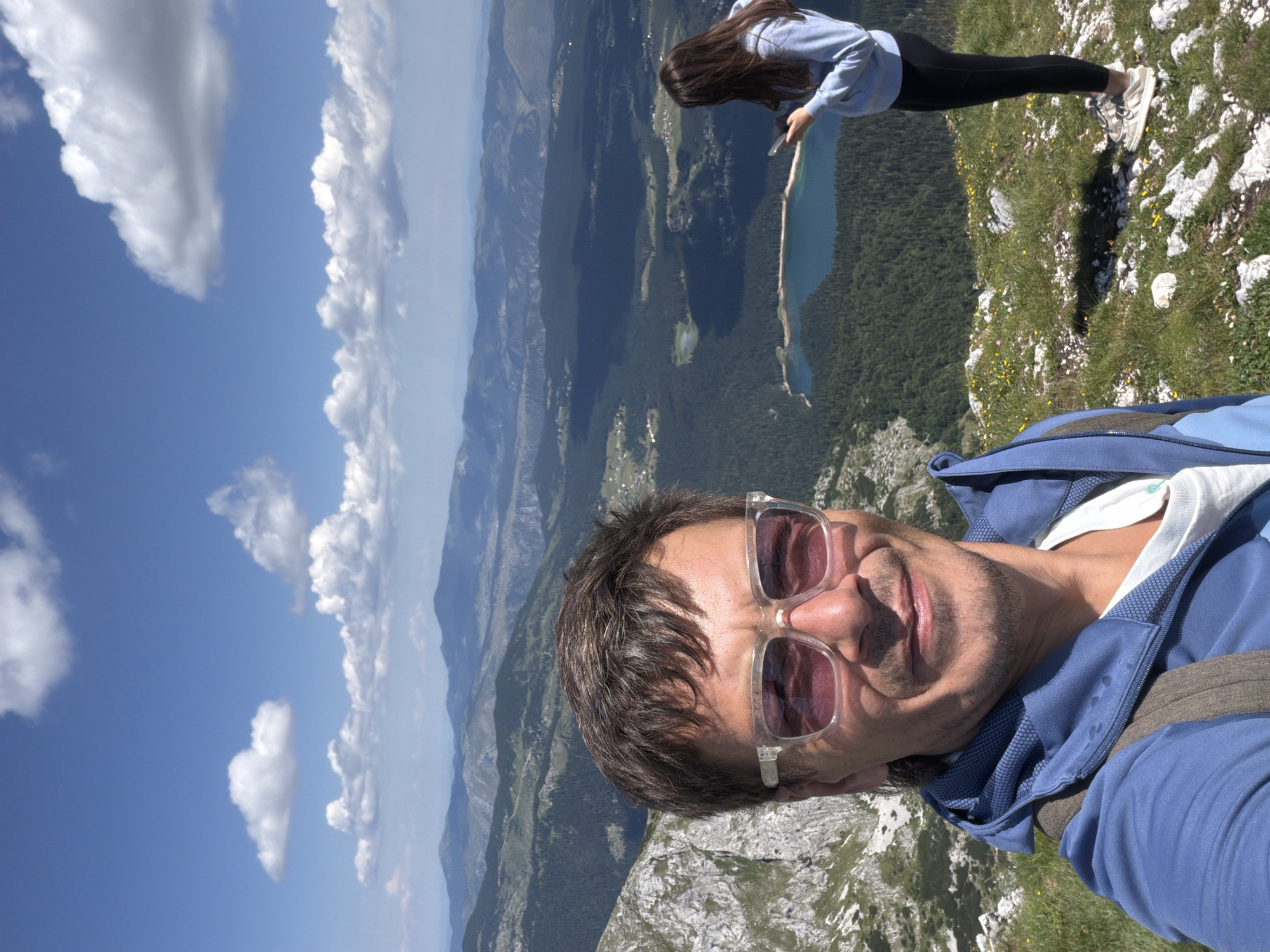 A selfie is taken by a man with short brown hair, wearing sunglasses and a blue hooded jacket, with a mountainous landscape in the background. To his right, a young woman with long brown hair is standing and looking at her phone, wearing a light blue hooded sweatshirt and black leggings. The landscape features a large lake, dense green forests, and rolling mountains under a blue sky with white clouds. The man appears to be in the foreground, while the woman and the landscape are in the background. The image is taken from a high vantage point, likely a mountain summit, overlooking a valley. The location is specified as Pašina voda, Montenegro.