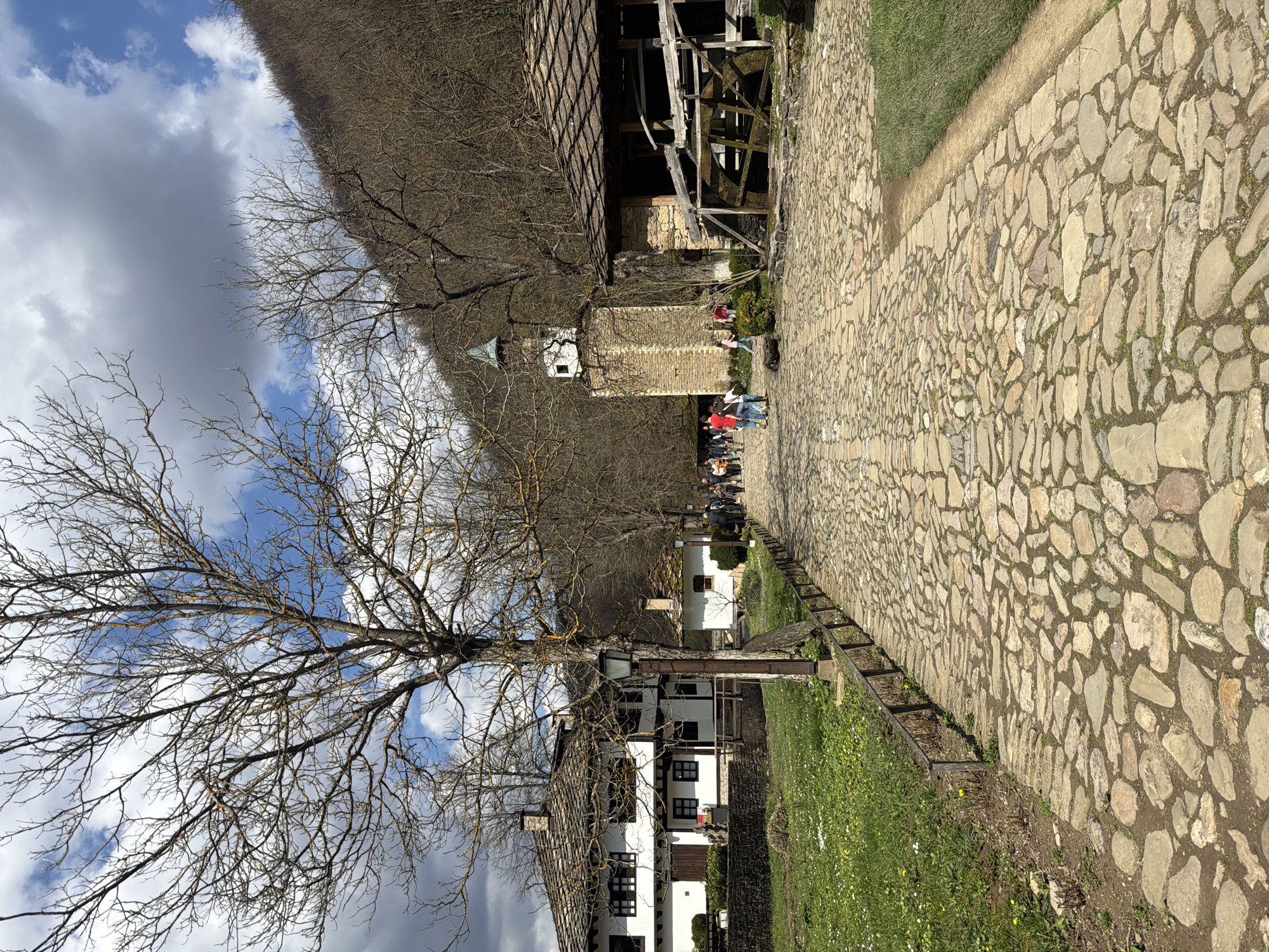 The image presents a view in Gabrovo, Bulgaria, featuring a stone-paved pathway lined with traditional buildings. Several people are walking along the path. A bare tree is on the left side of the image. Buildings with white walls and dark-tiled roofs are visible along the path. A wooden structure with a water wheel is visible. The sky is partly cloudy. There is green grass on either side of the pathway.