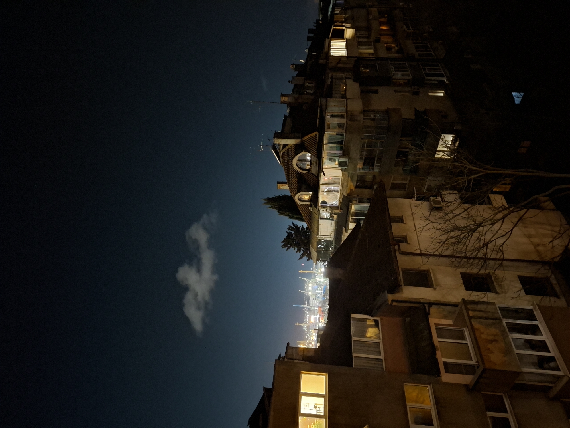 The image shows a nighttime scene in Burgas, Bulgaria. Buildings are the predominant feature, illuminated by internal lights. The sky is a deep blue, with a few clouds visible. A cityscape with bright lights is visible in the distance, possibly a port.