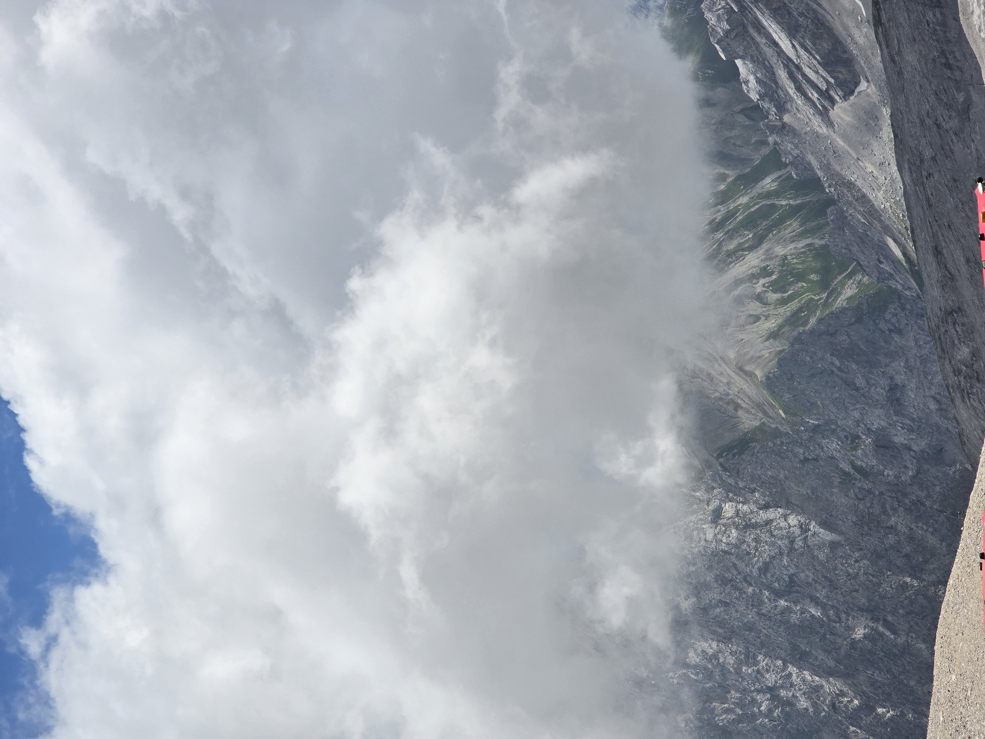 The image depicts a mountainous landscape with a dramatic sky. A large portion of the frame is occupied by thick, white clouds that partially obscure the rugged gray and green slopes of the mountains. The mountain terrain is steep and rocky, with patches of green vegetation visible in some areas. In the foreground, there is a gravelly surface, and the edge of a red structure, possibly a train or building, is visible at the very bottom of the image. The scene suggests a high-altitude location with variable weather conditions, characteristic of the Bavarian Alps. This view is likely from the Zugspitze, Germany's highest peak, located near Garmisch-Partenkirchen. There are no visible people in the image.