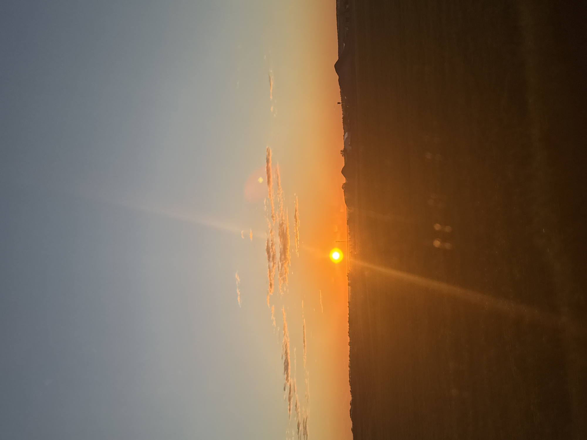 The image shows a sunset in Сыйрышбашево, Russia. The sun is near the horizon, casting a warm orange glow across the sky and landscape. There are some light clouds to the left of the sun. The land to the right is dark, obscuring details, but the silhouette of a town or village is visible along the horizon line.
