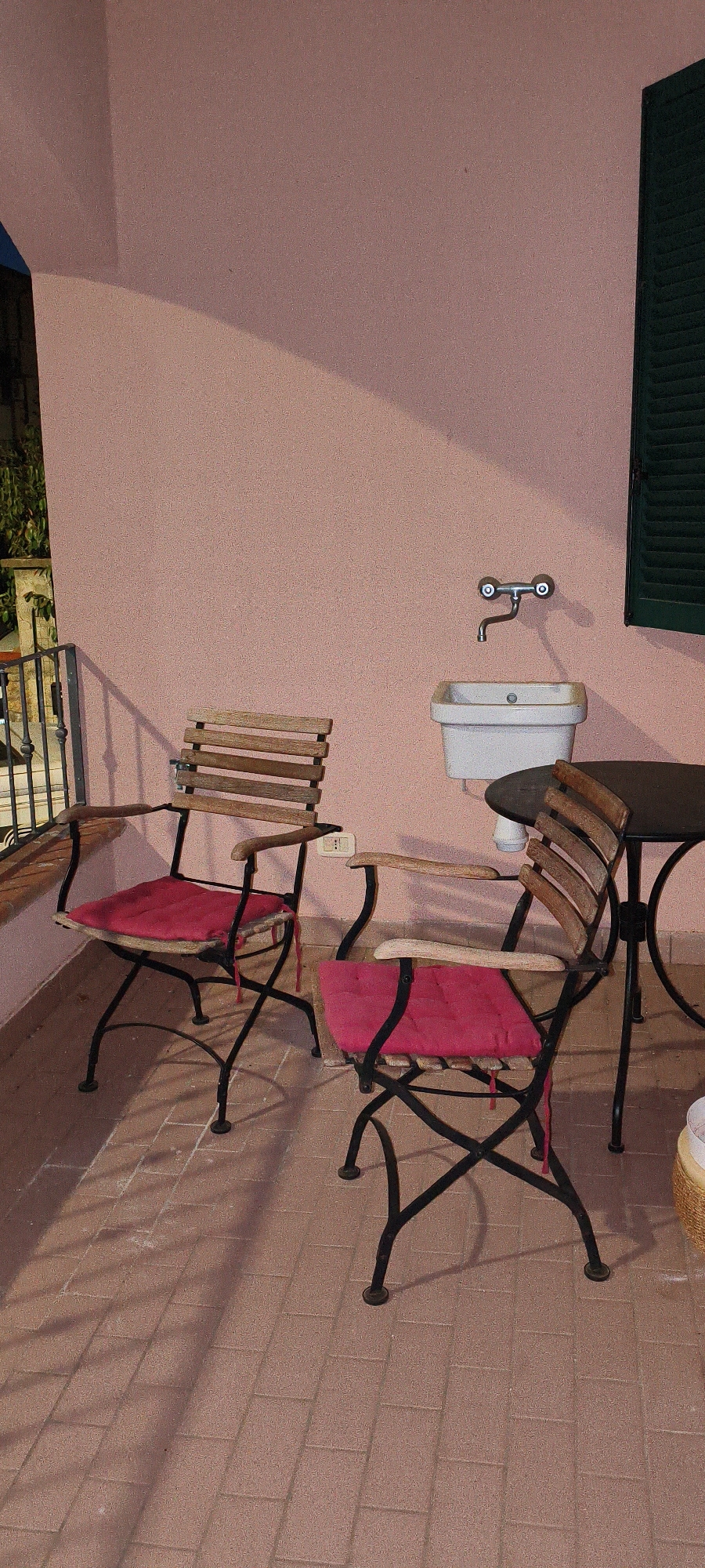 The image depicts an outdoor balcony or patio area, likely in Campagnatico, Italy. The wall of the building is painted a light pink color. On the balcony, there are two wooden chairs with black metal frames, each fitted with a bright pink cushion. In the background, a white sink with a silver faucet is mounted on the wall. To the right of the sink, a dark-colored round table with a black metal base is visible, with another wooden chair to its right. A dark green shuttered window is present on the far right of the image. The floor is paved with reddish-brown rectangular tiles. A metal railing is visible on the left side of the image, and some greenery can be seen beyond it. The lighting suggests it is either early morning or late afternoon, with shadows cast across the tiled floor.
