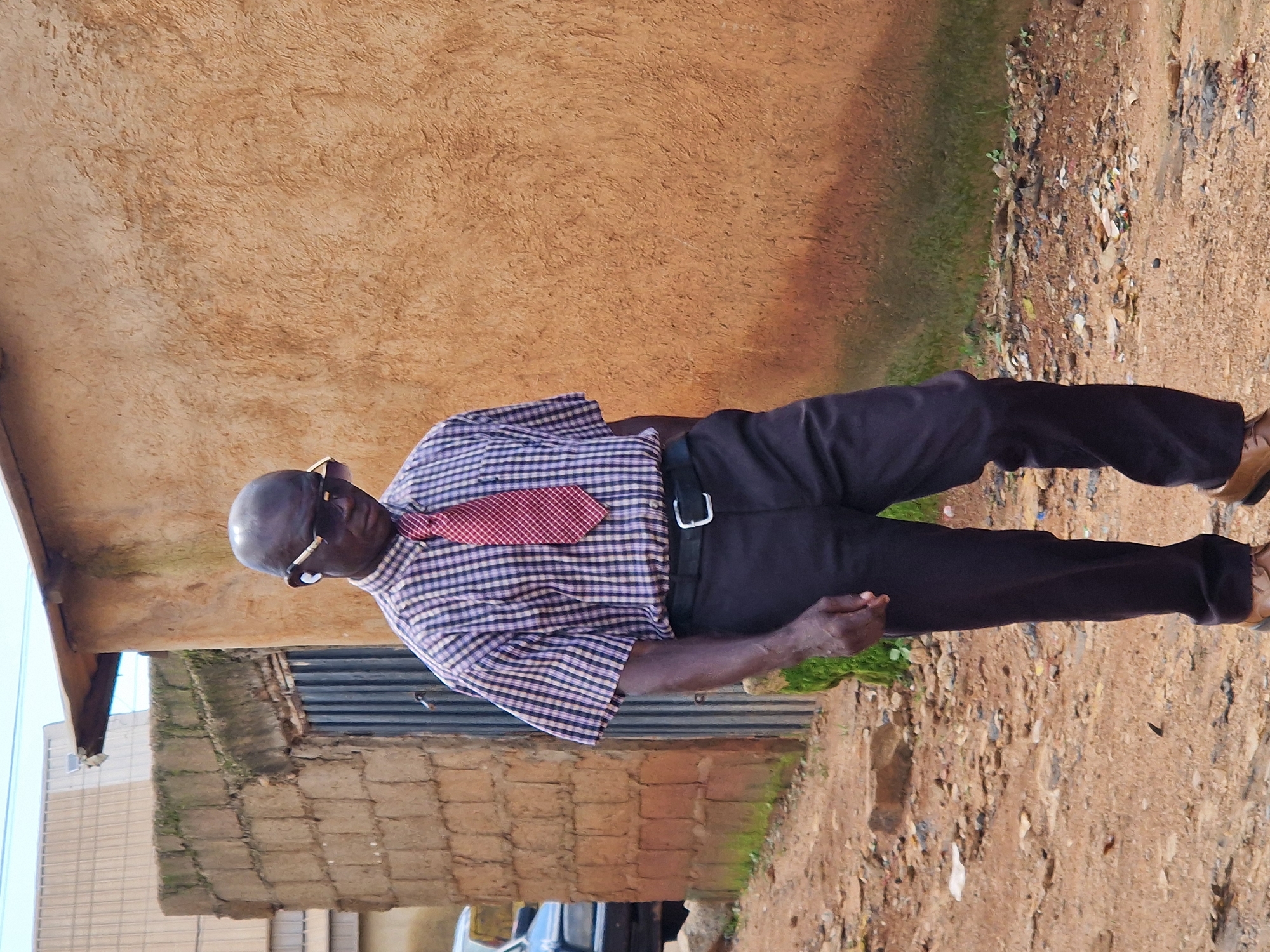 A full shot captures a dark-skinned man wearing sunglasses and a red and white checkered shirt with a red patterned tie, walking outside. He has short hair and is wearing dark pants and brown shoes. He is wearing white earbuds. The man is walking in front of a textured orange wall and a brick building. To the left of the man, a blue car is partially visible. The ground is dirt and gravel, with scattered debris.