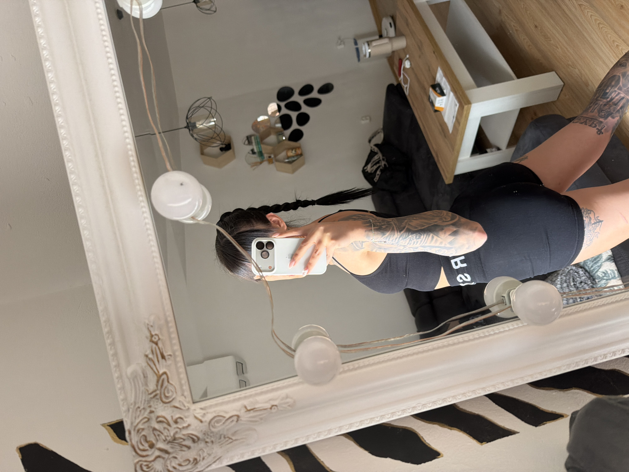 A woman is taking a mirror selfie in a room. She is wearing a black sports bra and black shorts. She has long dark hair in a braid and multiple tattoos on her arms and legs. The room appears to be a living room with a couch, a coffee table, and a large ornate mirror. The floor is made of wood. The woman is holding a phone in her right hand, which is obscured by her hair. The wall behind her has a striped pattern. There are several light fixtures and shelves on the wall, visible in the reflection. The scene is indoors.