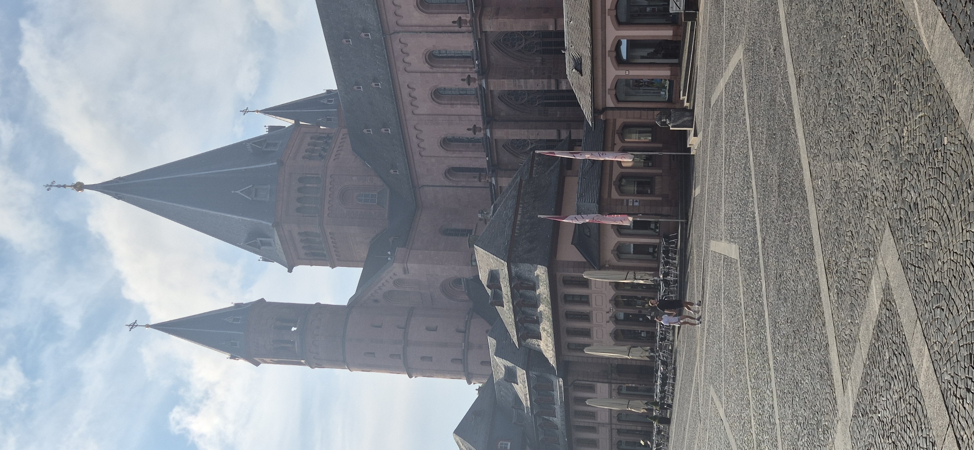 The image shows a view of the Mainz Cathedral (St. Martin's Cathedral) in Mainz, Germany. The cathedral is a large, historic Romanesque cathedral made of red sandstone, with multiple towers and spires. In front of the cathedral is a large, open plaza paved with cobblestones. Several people are walking across the plaza. On the right side of the plaza, there are cafes with outdoor seating, some of which are covered by large umbrellas. Flags are also visible in front of some of the buildings. The sky is blue with scattered clouds. The image appears to have been taken on a sunny day.