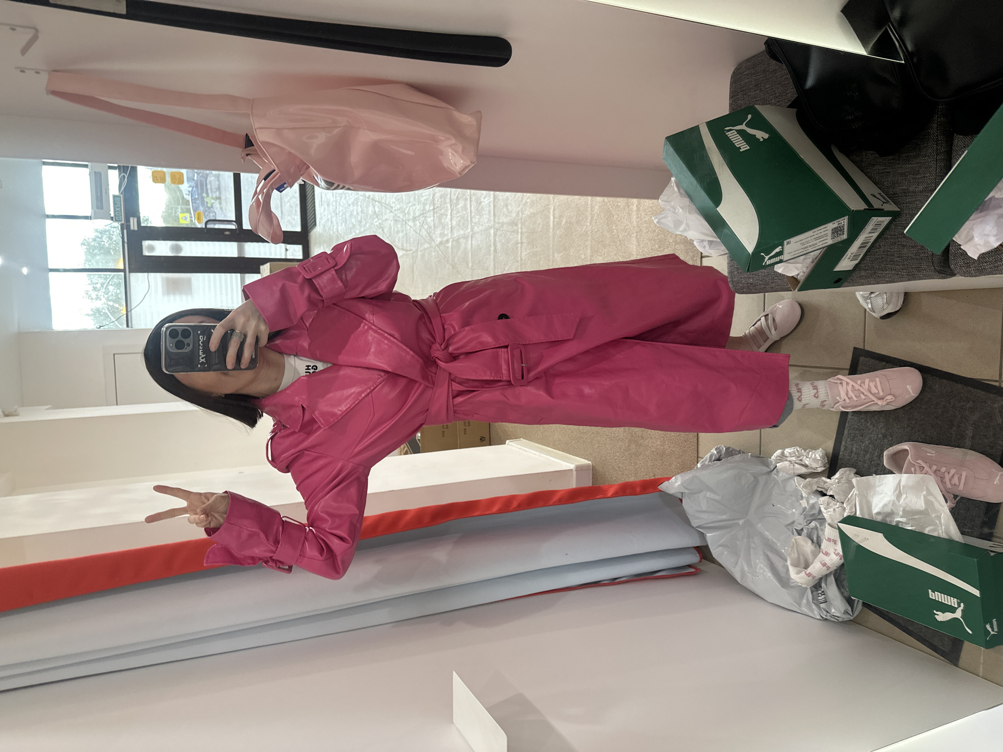 A person stands in a retail store environment, reflected in a mirror. The individual, holding a smartphone, wears a pink trench coat, pink socks, and matching sneakers. They display a "peace" sign with their hand. Visible objects include a pink bag, Puma shoeboxes, clothing items, and the store's interior features, such as shelves and display units. The location is a retail store, but the city of Votkinsk, Russia is not discernible from the given image.