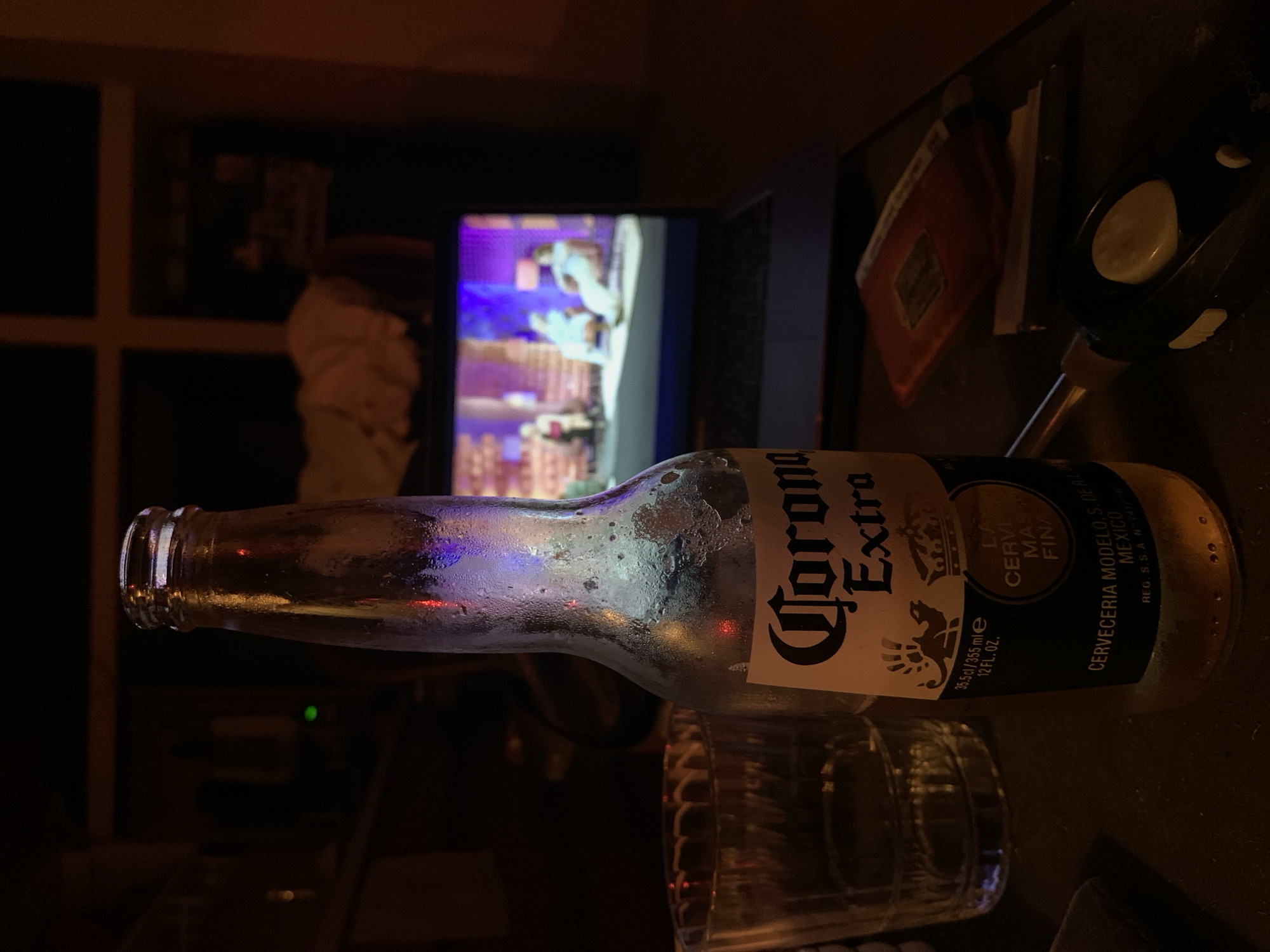 The image depicts a dimly lit indoor scene with a bottle of Corona Extra beer as the central focus. The bottle appears partially empty and condensation covers its surface. It is resting on or near a glass, which is partially visible. In the background, a laptop screen displays a scene with figures bathed in blue and white light. To the right of the laptop, a black object with a white button and a red rectangular item are visible. Behind the laptop, a window frame with a white cross-like pattern is discernible. The overall atmosphere is suggestive of a relaxed, casual setting. The location in Tel-Aviv, Israel, cannot be determined from the visual information provided.