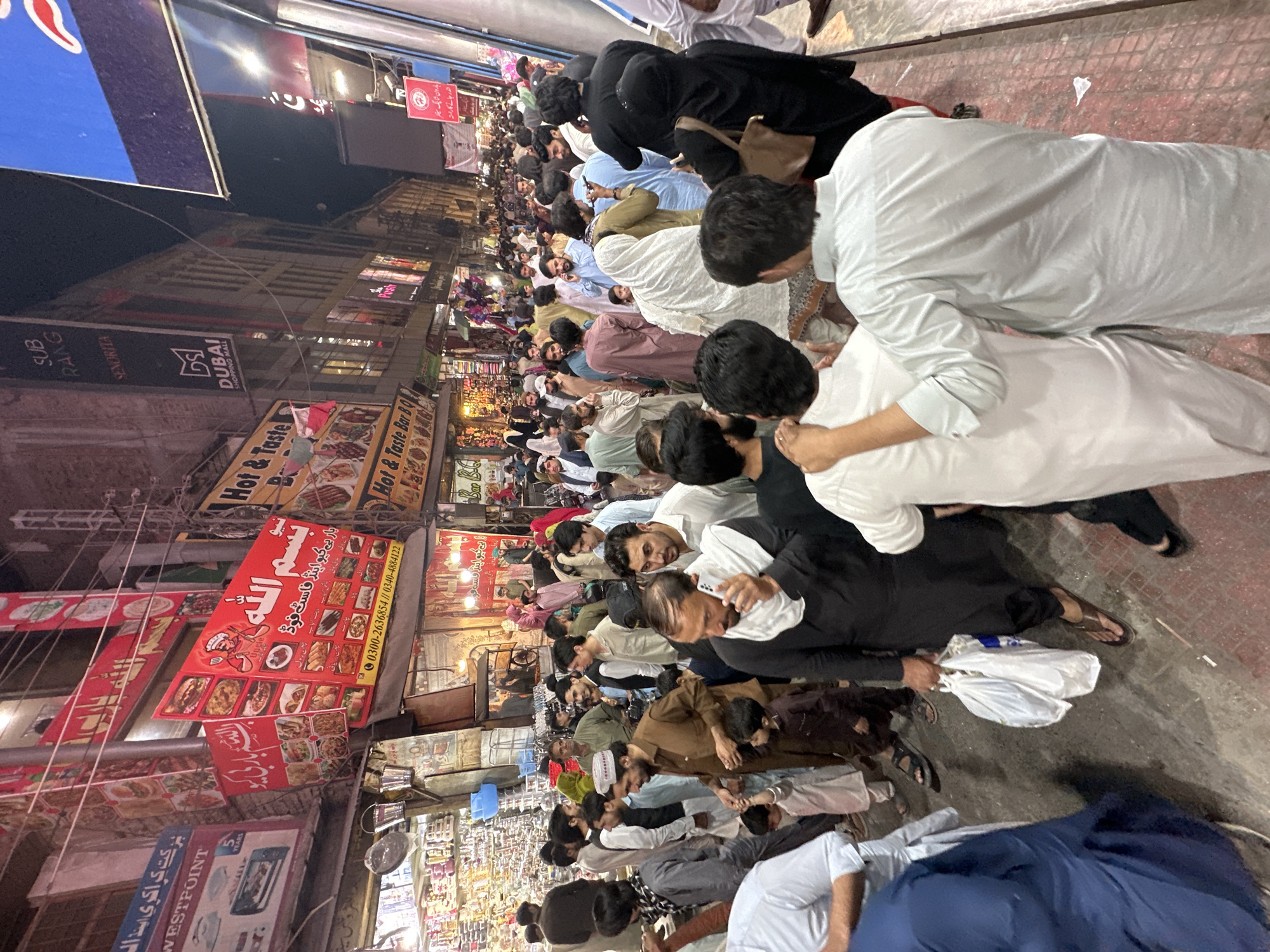 Here is a detailed description of the image:
**Scene Overview:**
The image shows a large group of people praying in a crowded, bustling urban street at night. The area is lined with shops and restaurants. The location is identifiable by local advertisements and signage in Urdu and mentions of "Quetta City Tehsil," suggesting it is a Pakistani setting.
**People:**
* The group consists mostly of men who are lined up closely together, seemingly performing a prayer.
* They are dressed in a mix of traditional Islamic clothing (thobes, shalwar kameez, head coverings) and casual attire.
* The men are arranged in rows facing the same direction, with some individuals assisting others.
* Some men are seen looking at their phones while praying.
**Environment and Objects:**
* **Street:** The street is narrow and paved with asphalt.
* **Shops:** Buildings lining the street house a variety of businesses, including restaurants ("Hot & Taste Bar BQ," " بار بی کیو اینڈ فاسٹ فوڈ"), and stores ("WESTPOINT," "DUBAI SHOPPING MALL").
* **Signage:** Numerous signs and posters adorn the buildings, advertising various products and services. The language is primarily Urdu.
* **Lighting:** The scene is illuminated by overhead streetlights and storefront lighting, creating a bright ambiance despite it being nighttime.
* **Objects:** A few plastic bags appear scattered on the ground near the praying individuals. Cables and wires crisscross overhead.
**Actions and Interactions:**
* **Prayer:** The primary action is the group prayer, with individuals bowing and kneeling.
* Some people are helping others stand straight.
* The proximity of the individuals suggests a shared sense of community and faith.
The scene depicts an everyday religious practice taking place in a busy commercial setting in Quetta City Tehsil, Pakistan.