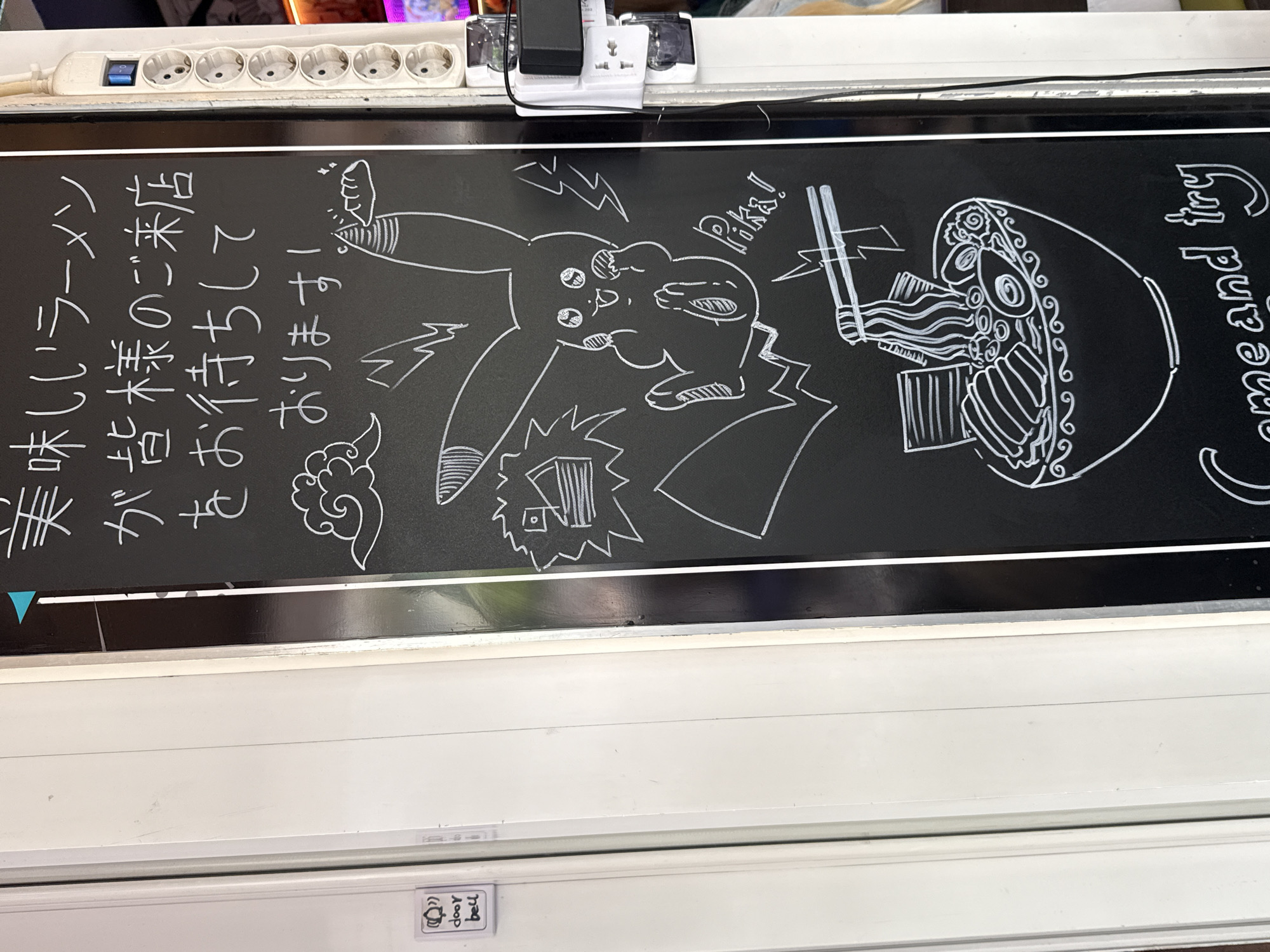 The image shows a chalkboard sign advertising ramen in Budapest, Hungary. The sign features a white chalk drawing of Pikachu holding chopsticks over a bowl of ramen with the text "Pika!" and "Come and try". There is also Japanese text. The sign is placed on a white surface near a power strip and a door bell sign.
