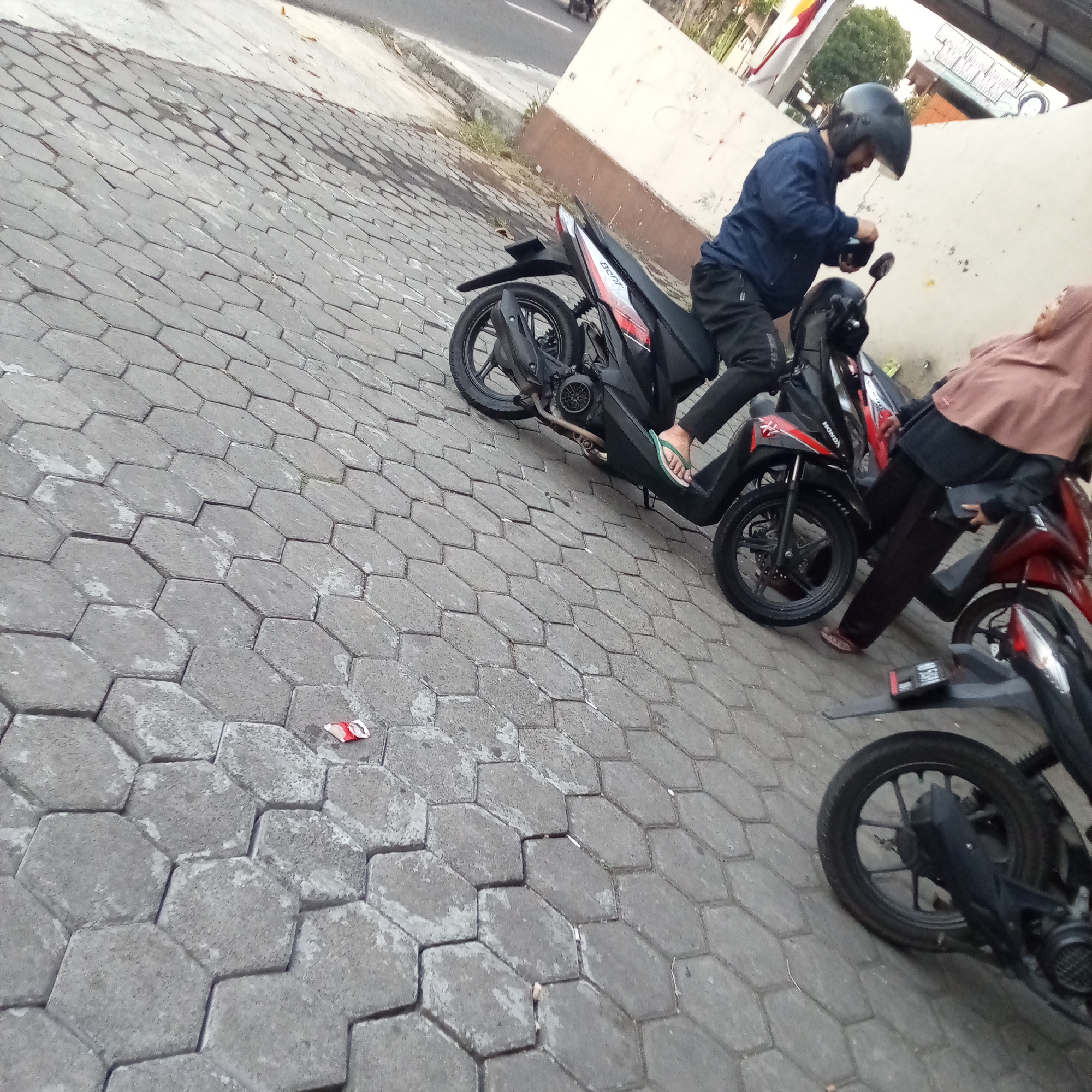 In Sidoharjo, Indonesia, a man wearing a blue jacket and a black helmet is seated on a black motorcycle, holding a black object. Beside him, a woman wearing a brown hijab and a dark top is seated on a red motorcycle. The motorcycles are parked on a hexagonal brick-paved street. In the background, a cream-colored building wall is visible, with some foliage and a glimpse of a road.