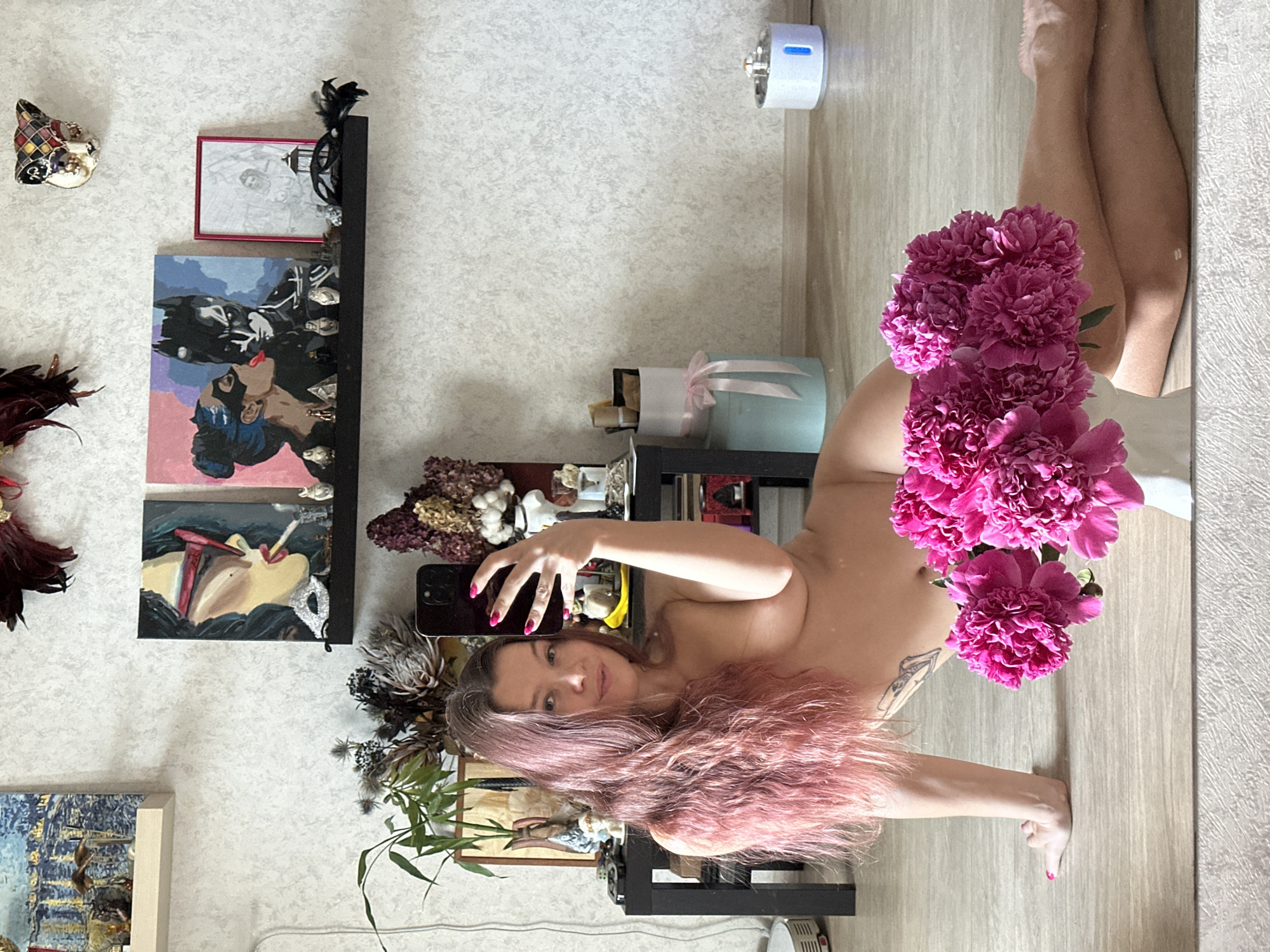 A nude woman with long pink hair is taking a selfie in a room. She is sitting on a wooden floor, propped up on her left arm. Her right arm is holding a smartphone, and she is looking at the camera. In front of her, partially obscuring her lower body, is a bouquet of bright pink peonies in a white vase. In the background, there is a dark rectangular table, various decorative objects on shelves, and a light-colored textured wall. The image also reflects a portion of the ceiling and floor, suggesting the photo was taken in front of a mirror. The location is not specified as Vladimir, Russia.