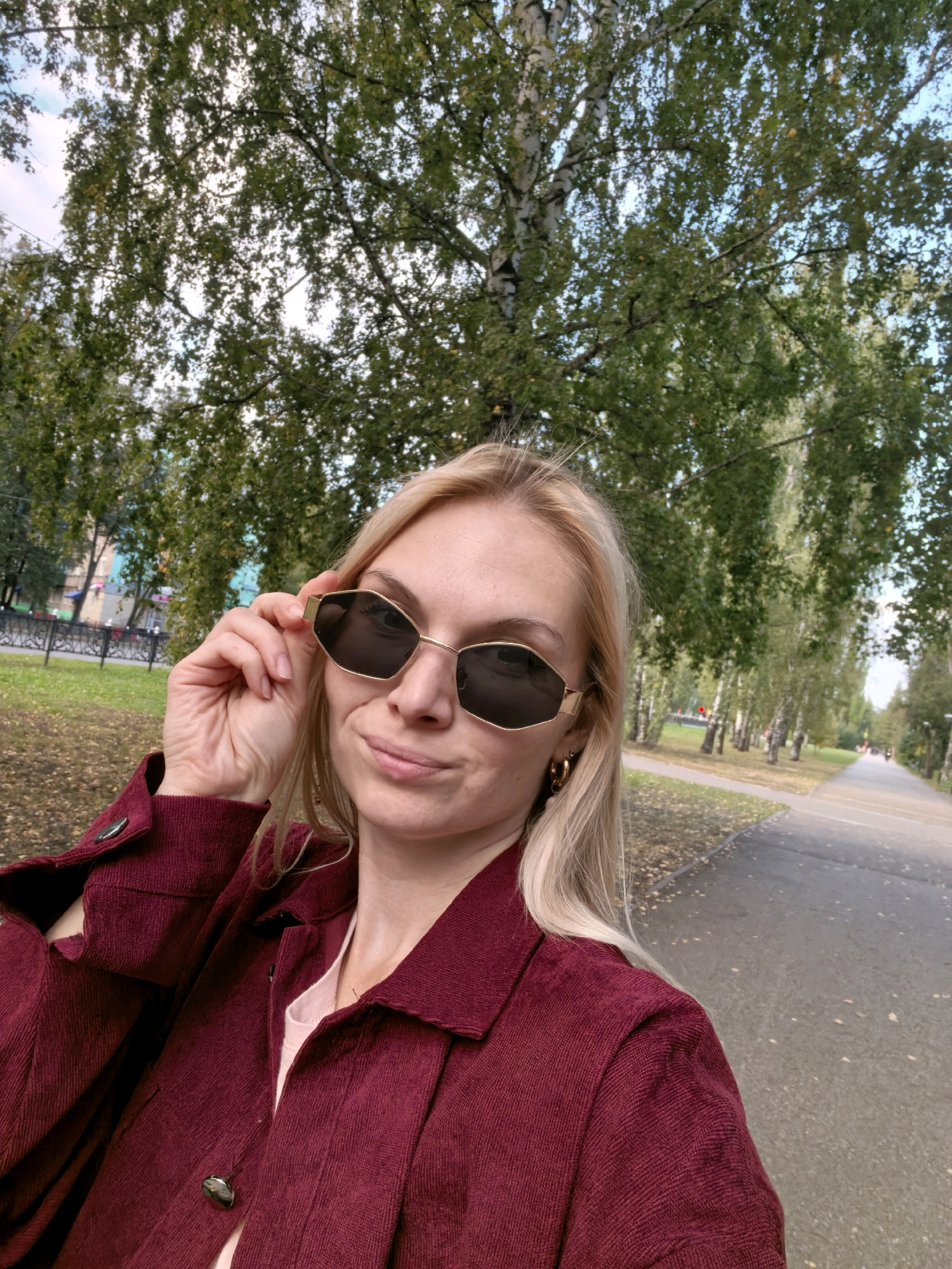 A blonde woman with sunglasses and a maroon jacket is taking a selfie outdoors in a park with trees and a pathway. The woman is adjusting her sunglasses. The pathway appears to be paved, and there are fallen leaves on the ground, suggesting it is autumn. The background shows a line of trees and some buildings in the distance. The image is taken during the daytime.