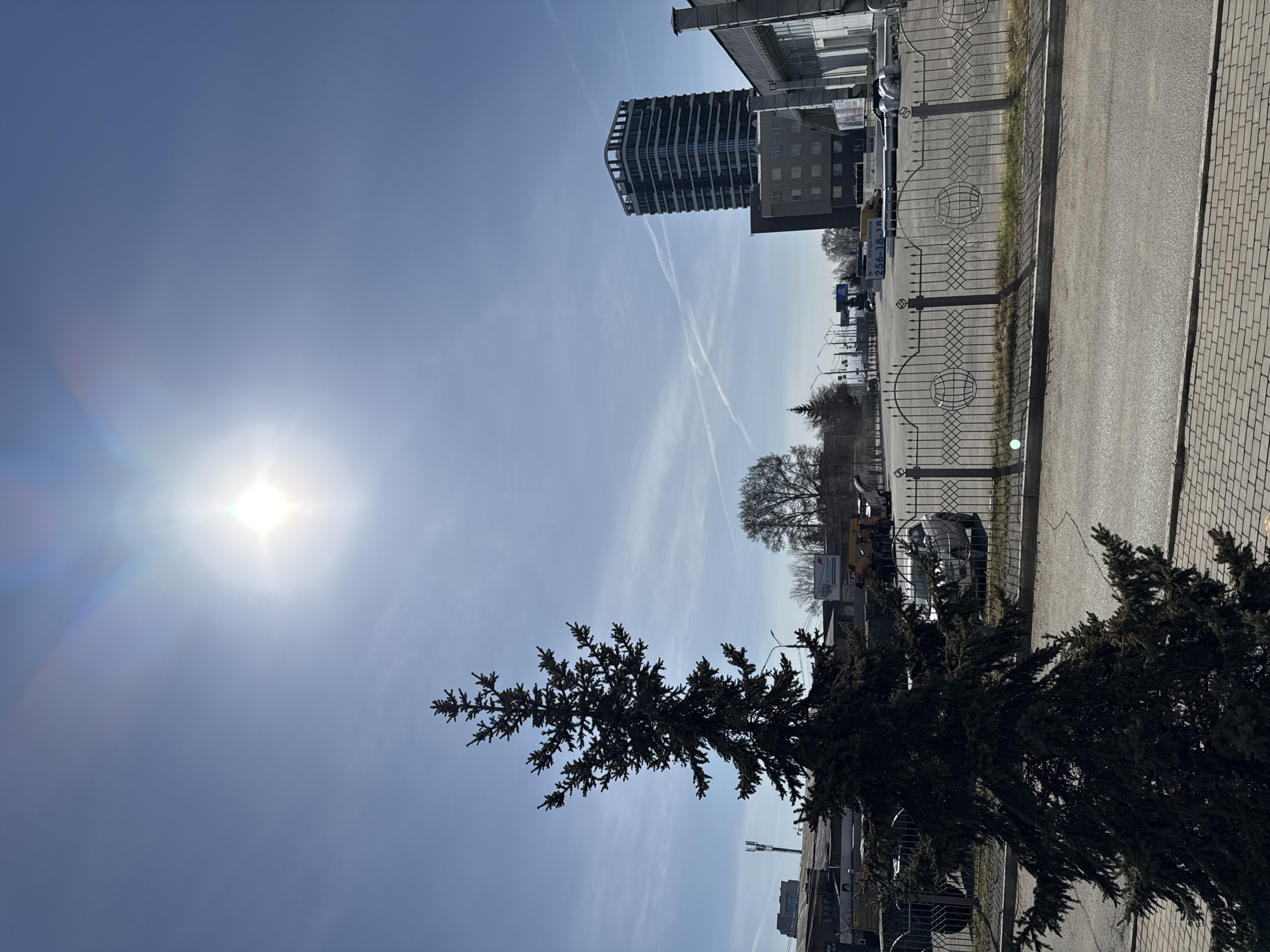 The image captures a sunlit street scene, oriented vertically, in Krasnoyarsk, Russia. The sun shines brightly in the upper-left quadrant, creating lens flare. Vapor trails crisscross the sky behind a tall modern building with a layered structure located slightly off-center. A decorative metal fence with circular designs borders a paved road, which transitions into a brick-paved area. A dark evergreen tree occupies the lower-right quadrant. Other buildings, trees, and vehicles are visible in the background, along with utility poles and street signs. The lighting suggests a clear day.