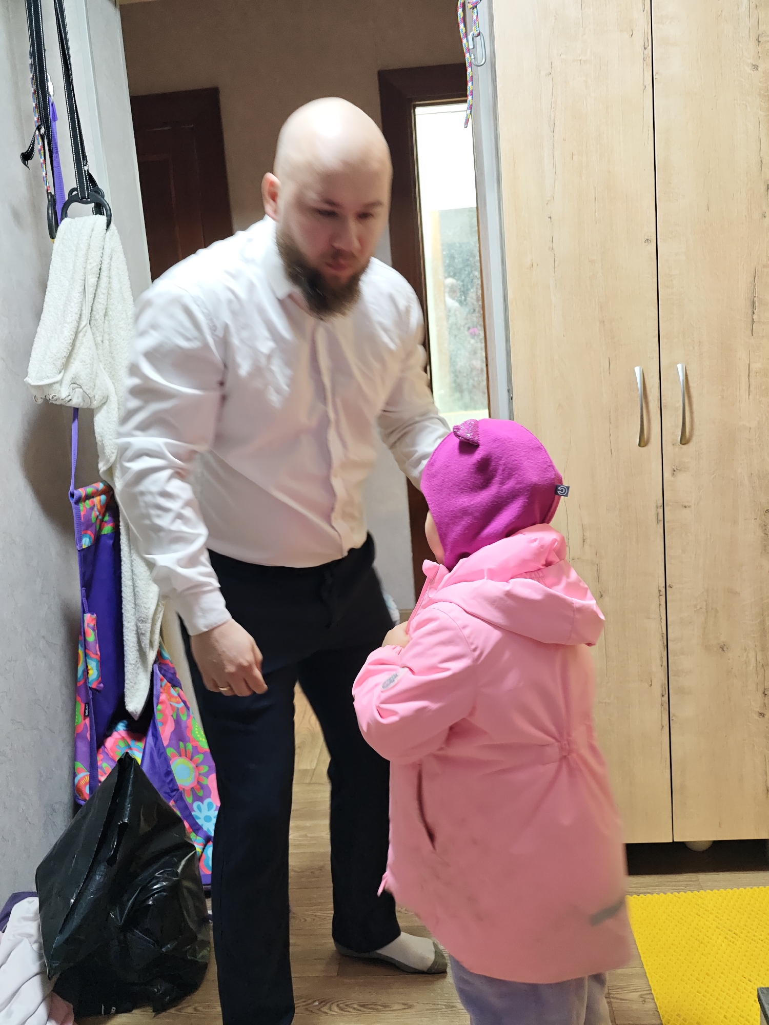 A medium shot depicts a bald, bearded man in a white shirt and black pants interacting with a young child in a pink jacket and purple hat. The man is looking down at the child. They are in what appears to be a hallway, with a closet to the right and a doorway visible behind the man. Items hang on the wall to the left of the man, including a white towel, and gym rings. A black bag rests on the floor. The photograph was taken in Chaikovsky, Russia.