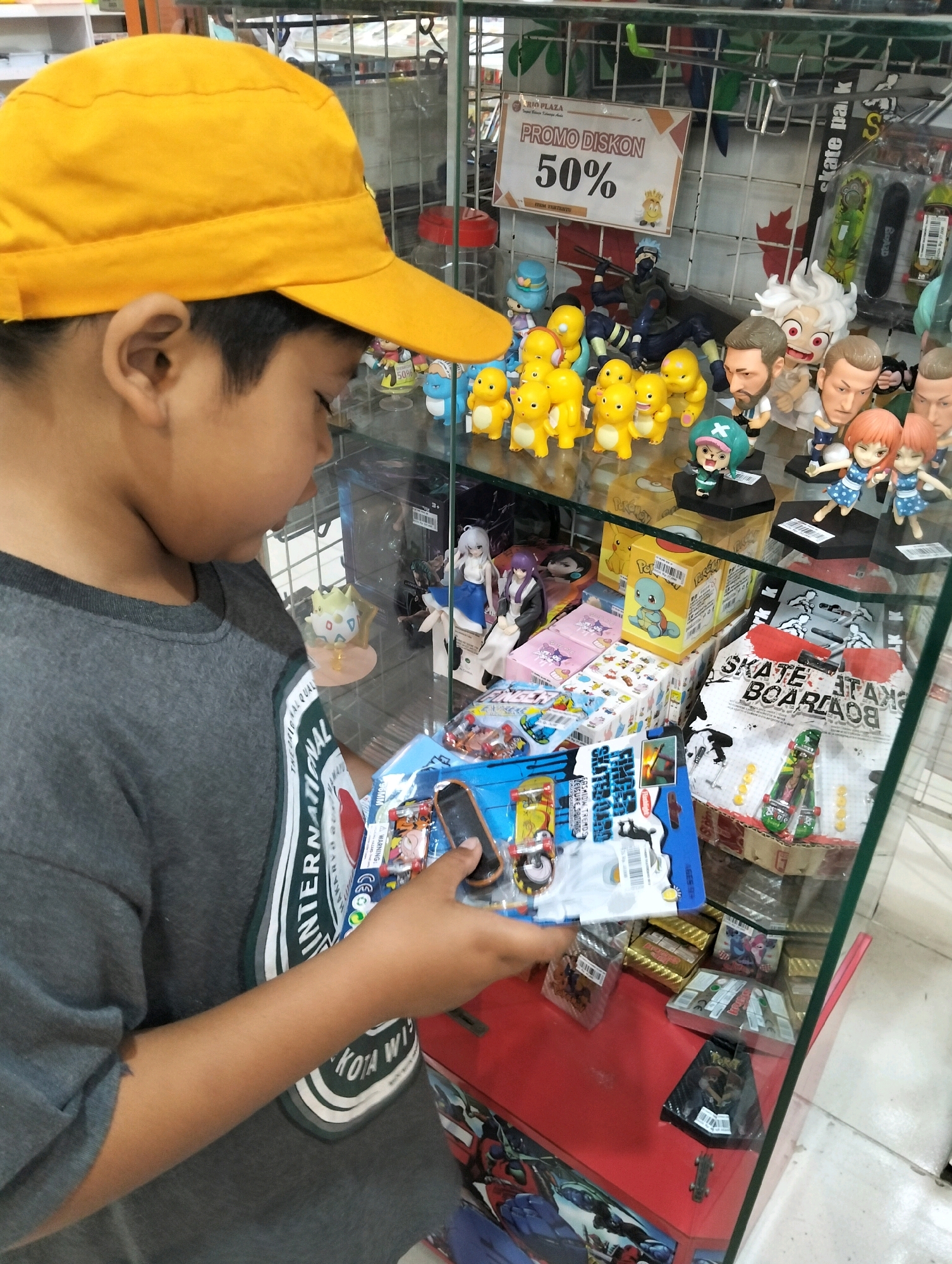 A young boy wearing a yellow cap and a grey t-shirt with a green and white striped design on the sleeve is looking at a display of toys in a store. He is holding a packaged toy in his hands. The display case contains numerous action figures, toys, and miniature skateboards. A sign in the background reads "PROMO DISKON 50%". The store appears to be a retail establishment selling collectible toys and novelty items.
