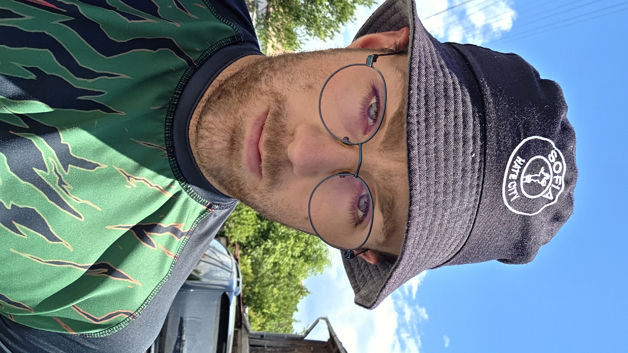 A close-up, eye-level outdoor portrait features a male subject, likely in his 20s or 30s, looking directly at the camera. He has light skin, a light brown beard and mustache, and is wearing round, thin-framed glasses with a subtle pinkish-purple lens tint. His eyes are visible through the lenses. A black bucket hat with a ribbed texture covers his head, featuring a circular white patch on the front. The patch reads "SOFIA" at the top, "HATE CITY" at the bottom, and depicts a stylized fist or hand symbol in the center. He is wearing a long-sleeved shirt with a dark grey/black collar and shoulder sections. The main body of the shirt is bright green with jagged, dark green/black abstract shapes outlined in a light tan color, resembling a camouflage or animal print. The background consists of a clear blue sky with sparse white clouds, green foliage from trees and bushes, and faint utility lines on the left. To the right, part of a dark wooden structure and a dark vehicle are visible. The scene is illuminated by direct sunlight, casting shadows. The location is in the city of Unknown, Unknown.