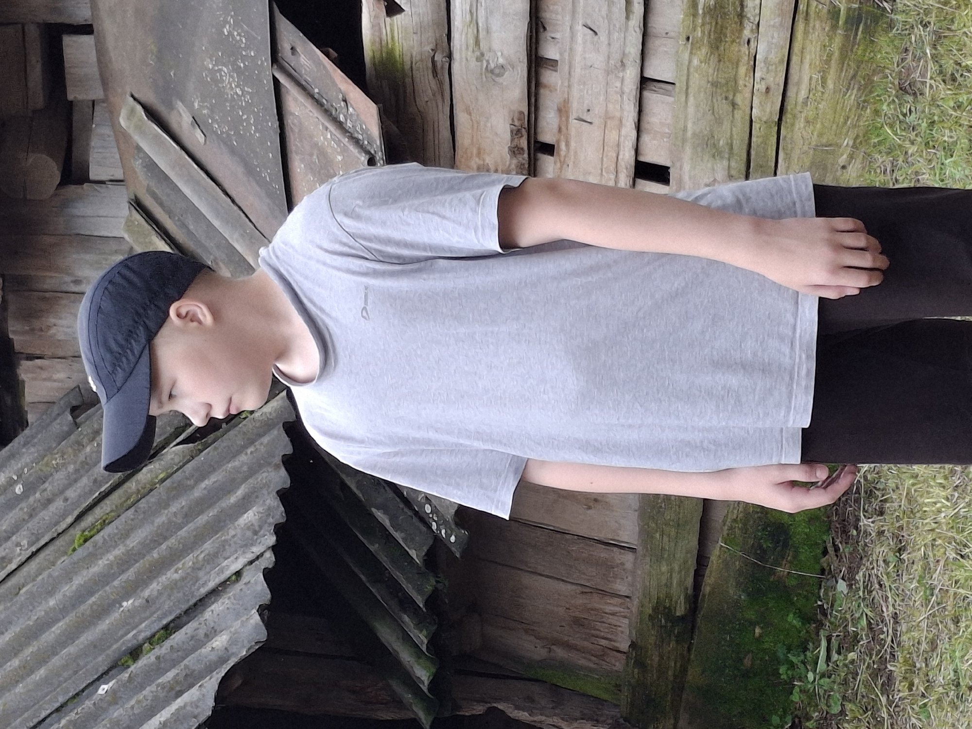A young boy wearing a grey t-shirt and a dark blue baseball cap is standing outdoors. He is looking downwards, his head tilted slightly. His left arm is bent at the elbow, with his hand resting on his lap. His right hand is also bent at the elbow and his fingers are slightly curled. The boy is standing in front of a weathered wooden structure, which appears to be a shed or an old building. Part of a corrugated metal roof is visible to the left of the boy. The background consists of more wooden planks and some green grass and foliage. The lighting suggests it is daytime, with diffused light indicating it might be overcast or late afternoon. The overall scene has a rustic and perhaps abandoned feel, consistent with a rural setting in Plishkino, Russia. No visible text is present in the image.