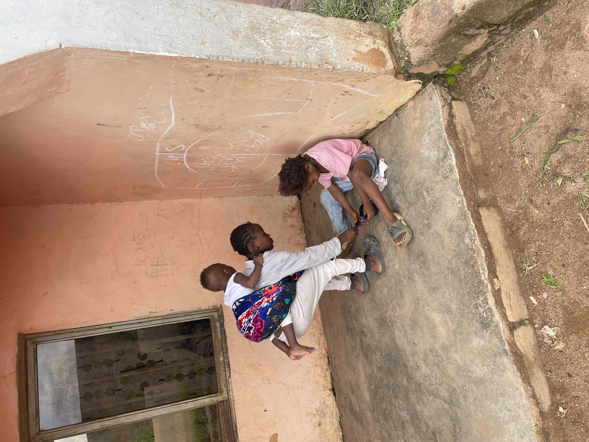 Three children are in an outdoor courtyard in Barakin Lerawi, Nigeria. One child, wearing a pink striped t-shirt and denim shorts, sits on the ground drawing on the wall with chalk. Another child, carrying a baby on their back, is also on the ground. The baby is wrapped in a colorful cloth. The third child appears to be sitting or kneeling nearby, out of frame. The courtyard has a concrete floor and a pale peach-colored wall with chalk drawings. A window with a metal grate is visible on the left. The scene suggests a casual daytime activity, possibly a break from school or a moment of play. The presence of chalk drawings and the simple setting indicate a likely rural or semi-rural environment.