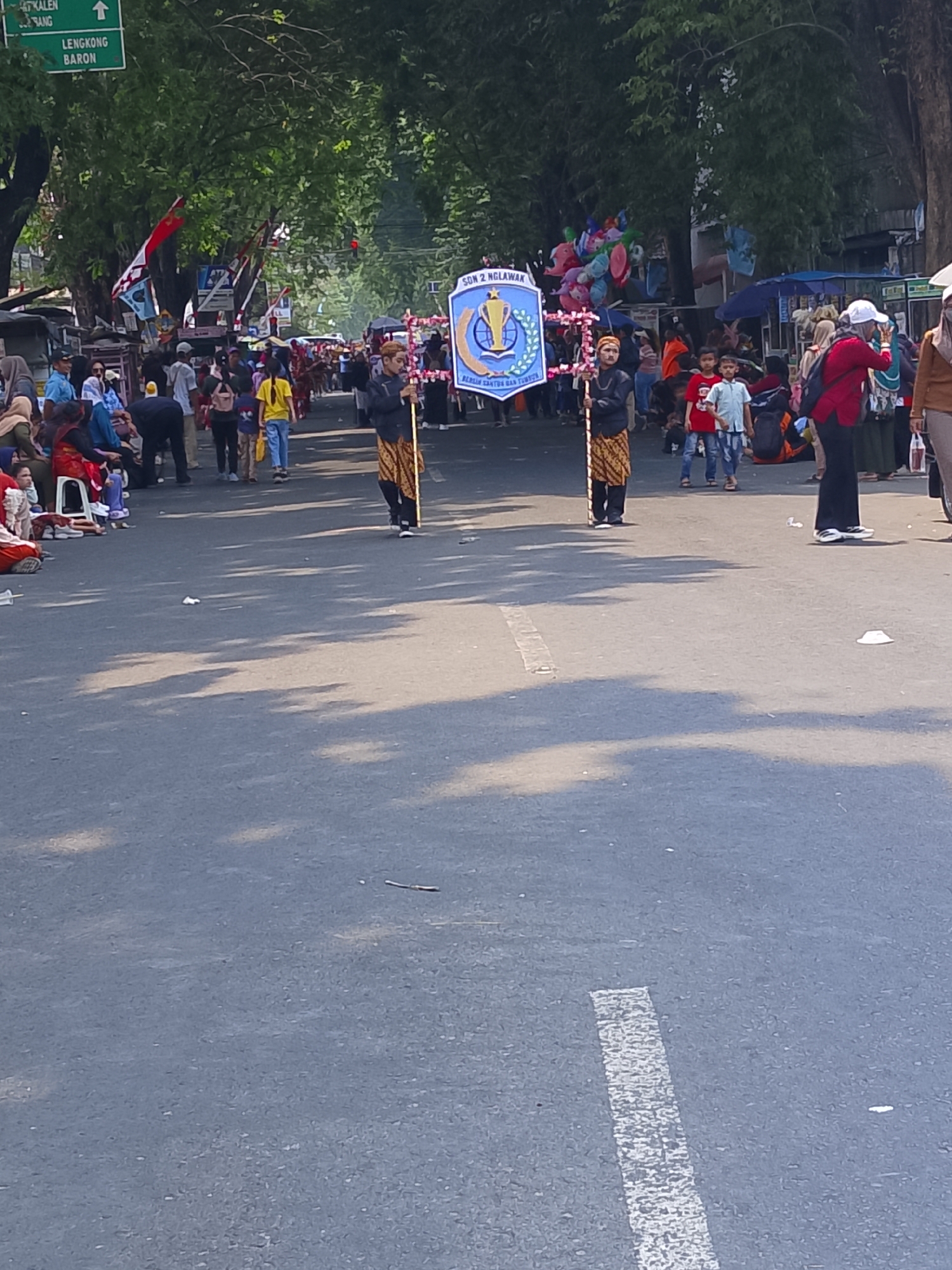 The image depicts a street scene in Kertosono, Indonesia, likely during a parade or cultural event. Two individuals, dressed in traditional attire including batik sarongs and head coverings, are holding a banner. The banner displays the text "SDN 2 NGLAWAK" and a crest with a trophy and circular emblem, indicating it represents a school. In the background, a crowd of people are gathered on the sides of the street, some seated and others standing. Children are visible among the spectators, and some are holding balloons. Vendors appear to be present, with stalls and tents lining the street. Trees with lush foliage form a canopy over the road, and sunlight casts shadows on the asphalt. A green street sign with white text indicates directions to "KALER," "LANG," and "LENGKONG BARON." The overall atmosphere suggests a festive and communal gathering.