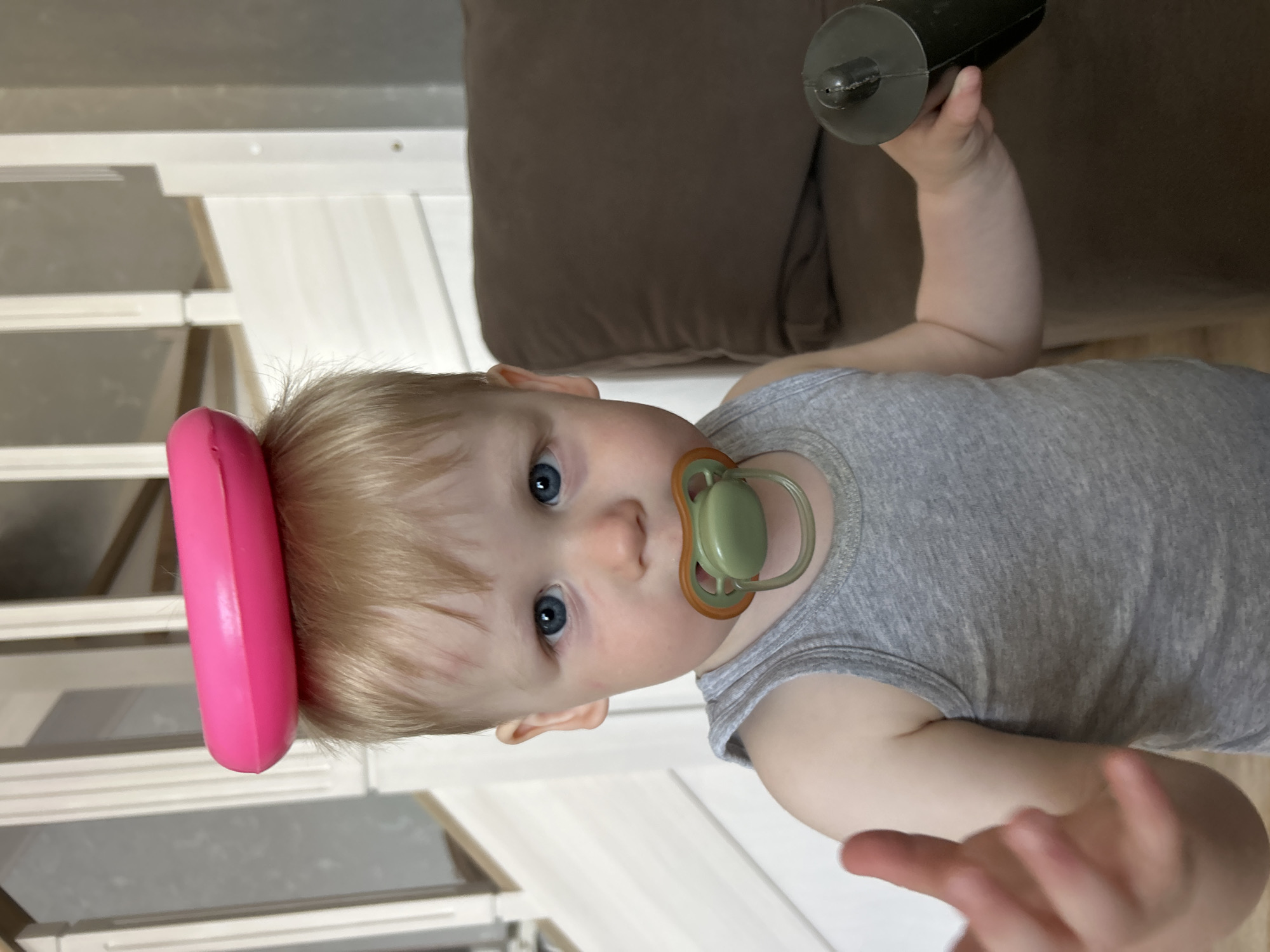 A young Caucasian child with blonde hair, blue eyes, and a pacifier in their mouth stands in a room. A pink ring is balanced on the child's head. The child is wearing a grey tank top and holding a black object in their right hand. The background shows a staircase with white banisters and a brown couch. The specific location in Chaikovsky, Russia cannot be determined from the image.
