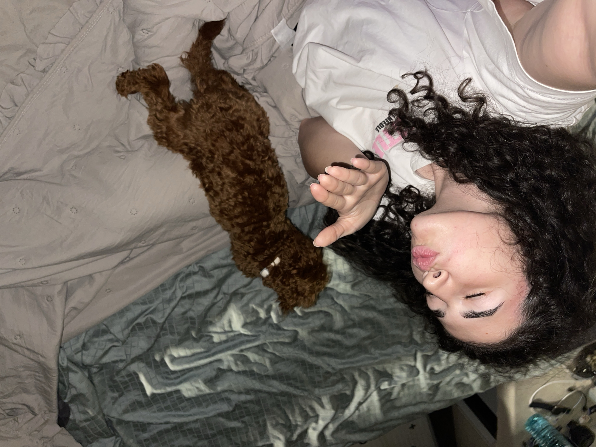 A high-angle indoor photograph depicts a person and their dog resting on a bed, likely in Sofia, Bulgaria. A female individual with long, dark, curly hair lies on her back on a light grey, textured duvet, her head positioned towards the lower right of the frame. Her eyes are closed, and her lips are softly puckered in a relaxed expression. She wears a white t-shirt, with the word "NEEDLE" partially visible on the fabric near her shoulder. Her left arm is bent, with her hand gently lifted.
Beside her, a small to medium-sized dog with dense, curly, reddish-brown fur, possibly a poodle mix, is stretched out on its side. The dog appears to be resting or asleep next to its owner. The bedding consists of a light grey ruffled duvet and a darker grey or teal blanket with a subtle grid-like pattern, which is visible beneath the duvet. The overall scene conveys a sense of peace and intimacy, suggesting a quiet moment of rest, possibly in the early morning or late evening. In the lower right corner, a few blurry items, including a blue water bottle and electronic cables, are visible on a surface next to the bed.