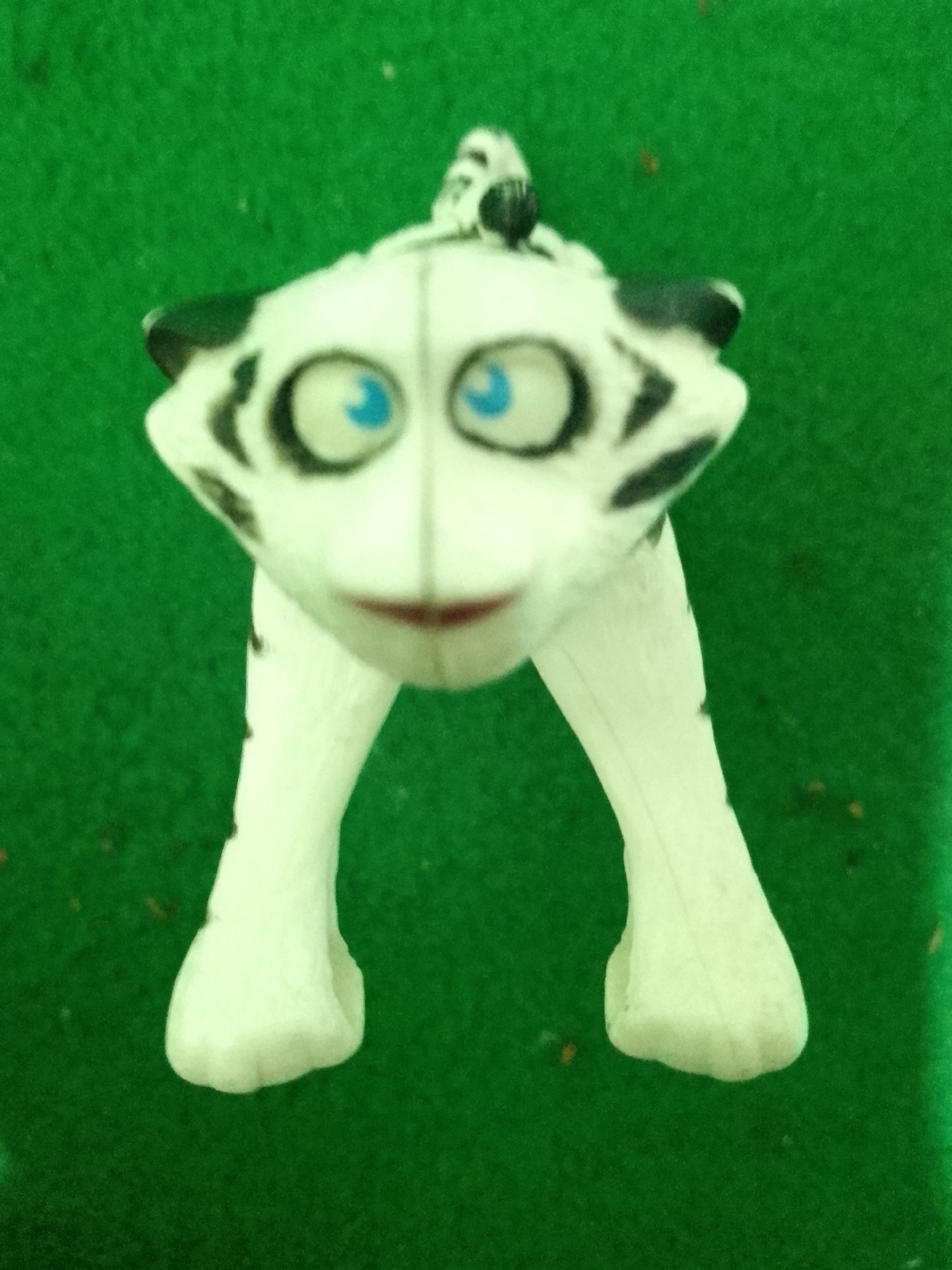A white, stylized toy tiger with blue eyes and black stripes is posed standing on a green, fuzzy surface. The tiger's head is tilted slightly forward, and its mouth is open, revealing a red interior. The toy is centrally located in the frame, with the green surface extending outwards in all directions. There is no visible indication of the city of Kauman, Indonesia in the image.