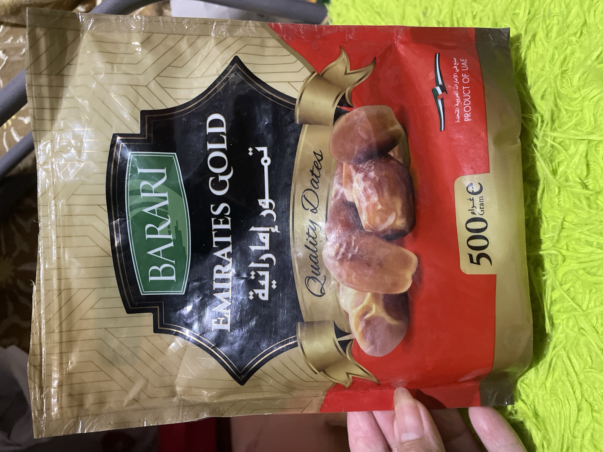 A slightly wrinkled package of "Barari Emirates Gold" dates rests on an undefined surface. The packaging is primarily gold-colored with a black central design element. The brand name "BARARI" is displayed in green, while the text "EMIRATES GOLD" and its Arabic equivalent "تمور إماراتية" are in black. Dates are depicted in a stack on a red section of the package, accompanied by text reading "Quality Dates." The package also indicates "PRODUCT OF UAE" and a weight of "500g". A human hand appears at the bottom of the frame. The scene is not identifiable as being in Davao City, Philippines, based solely on the provided image.