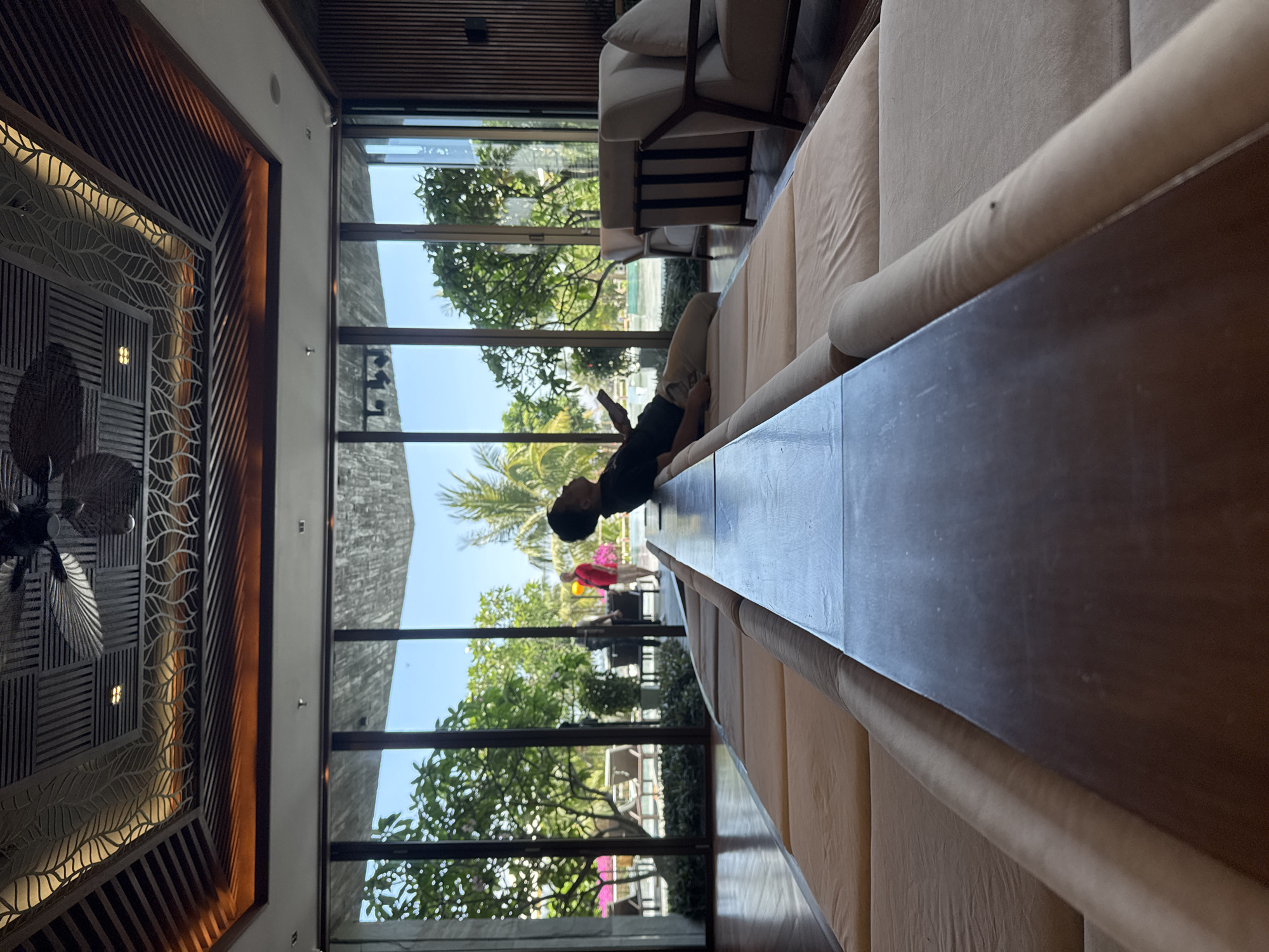 A medium shot captures a person reclining on an indoor sofa with floor-to-ceiling windows showcasing a lush outdoor landscape. The image appears to be rotated counter-clockwise to 90 degrees, with the ceiling featuring intricate, dark-wood detailing contrasted against lighter, patterned elements. Outside the window, greenery and a glimpse of a sunny sky suggest a tropical climate. The person, positioned towards the center of the frame, is holding a phone. This image was taken in Đà Nẵng, Vietnam.